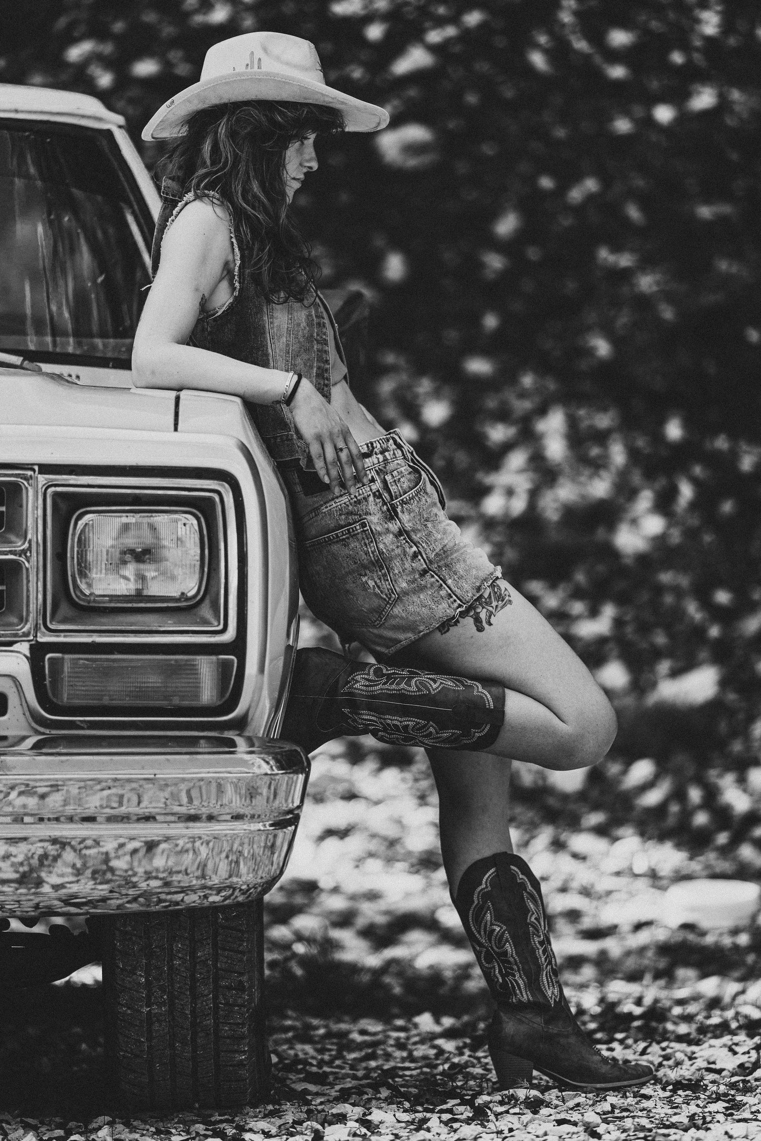 Black and white photo of a woman in cowboy hat, denim overalls, and boots leaning against a vintage pickup truck.