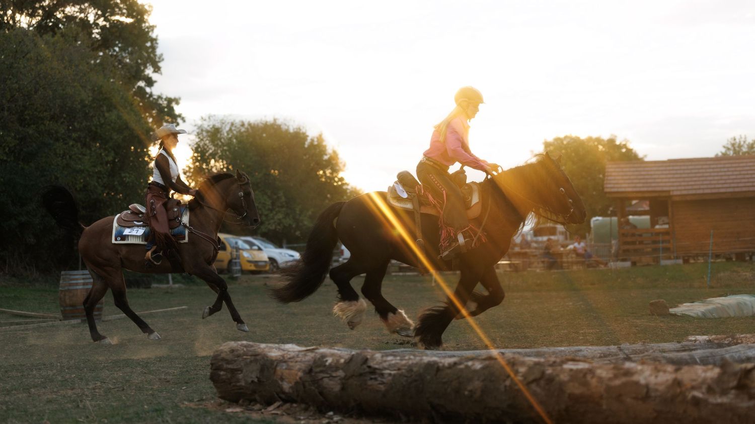 Two riders on horseback jumping over logs in golden sunset light at an outdoor equestrian course.