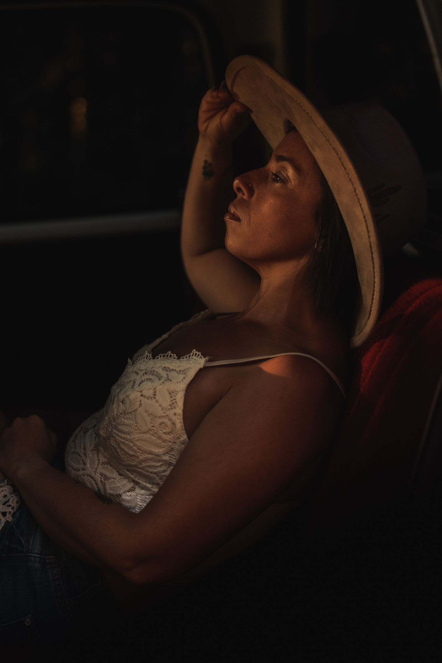 Woman in white lace top and straw hat relaxing in warm dramatic light, looking upward with a contemplative expression.