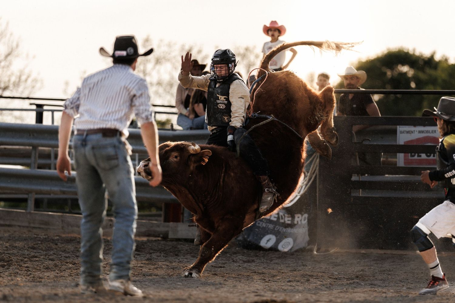 A cowboy rides a bucking bull at a rodeo while a rodeo clown stands nearby in the dusty arena.