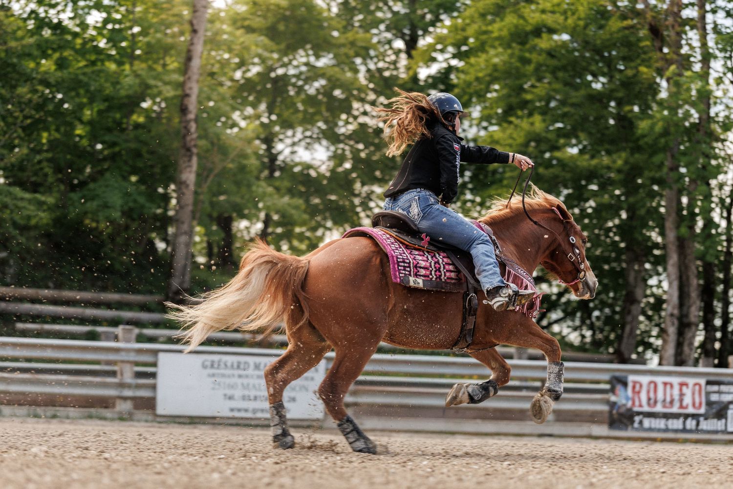 Woman riding a chestnut horse at a rodeo arena, wearing a black hat and jacket on a sandy track.