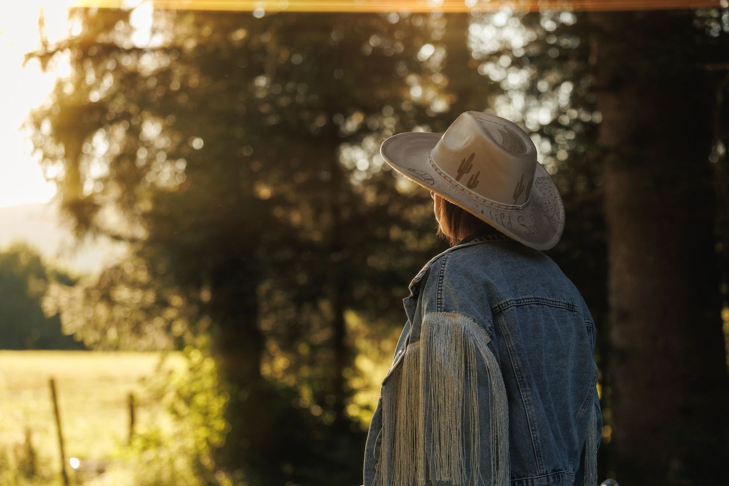 Woman in denim jacket and straw hat gazes at golden sunset light filtering through tall trees in a rural field.