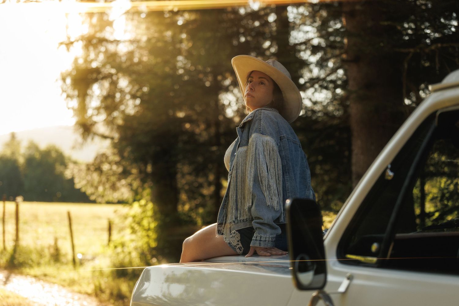 Woman in cowboy hat and denim jacket sitting on white truck hood in golden sunlight near green trees.