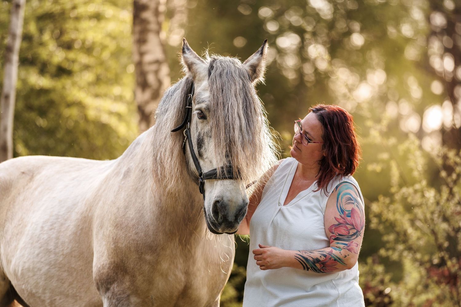 A tattooed woman in a white dress stands beside a grey horse in a sunlit forest setting.