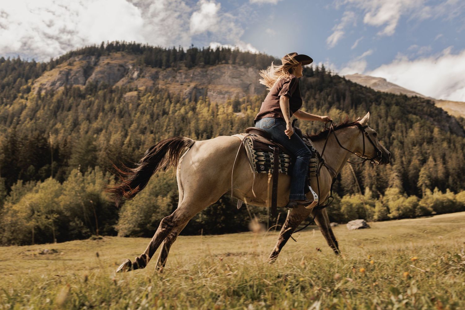 Woman in cowboy hat riding a galloping horse through a scenic mountain meadow with autumn trees.