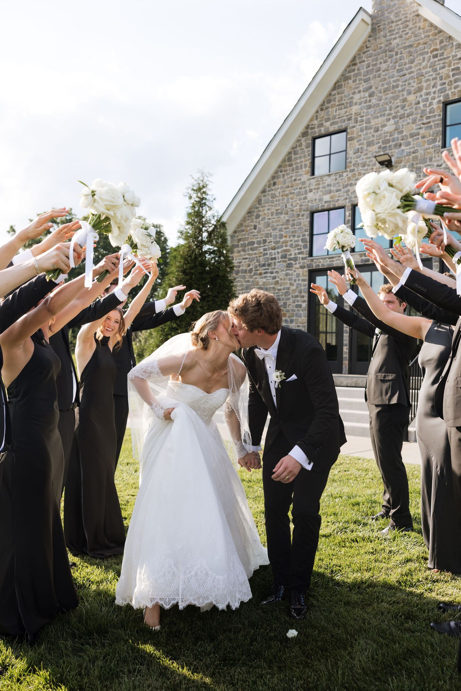 Bride and groom kissing under arch of raised bouquets formed by wedding party outside stone venue.