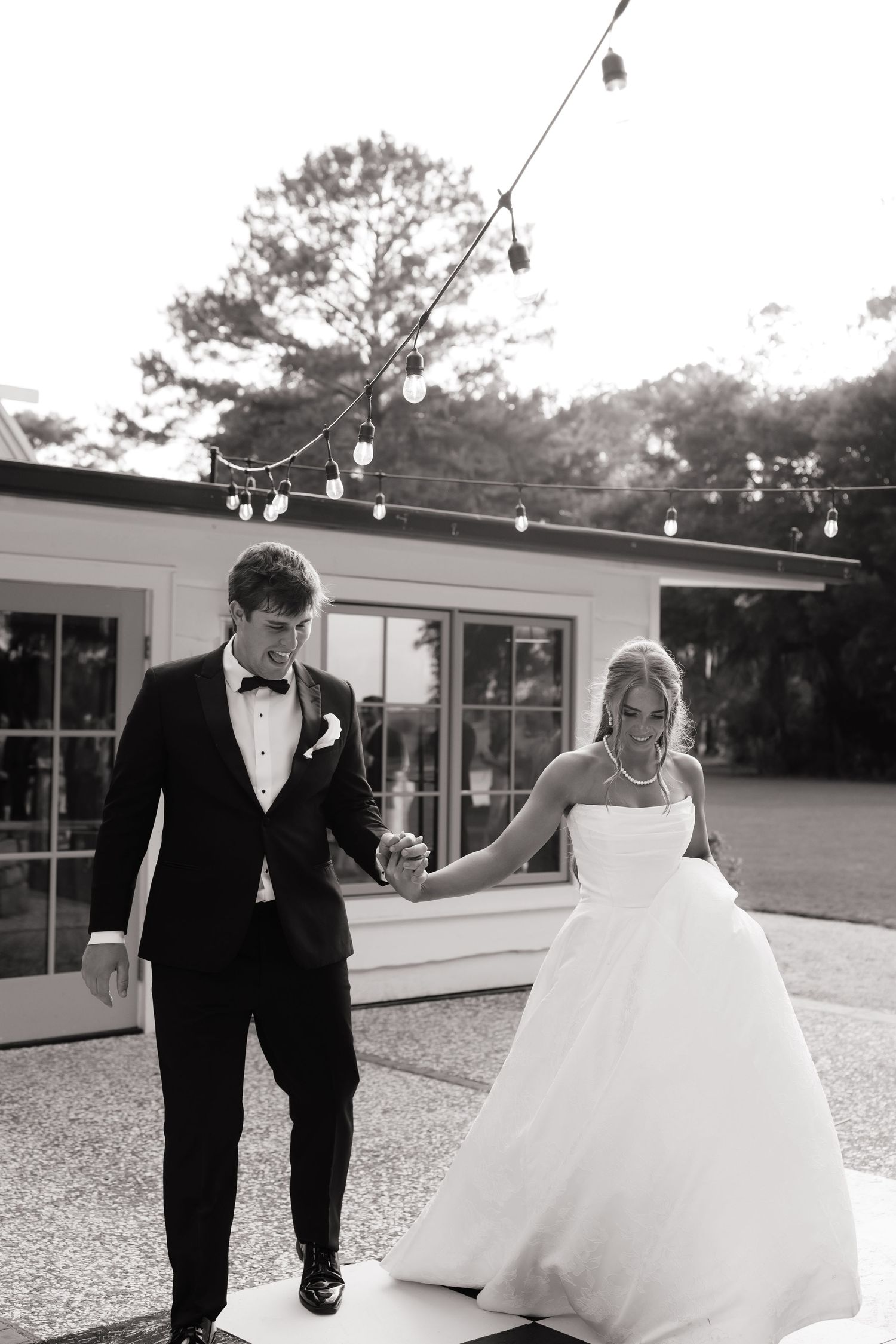 Black and white photo of a bride and groom holding hands, walking outdoors with string lights and a building in the background.
