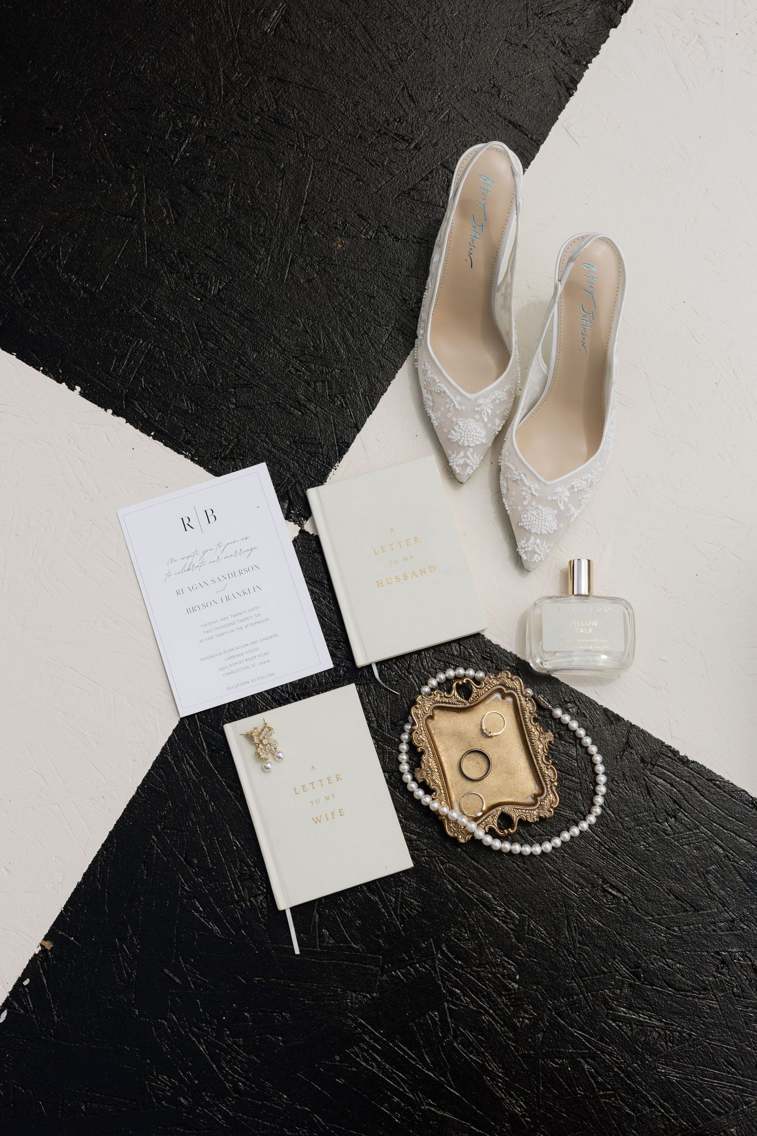 Elegant wedding flat lay with lace heels, pearl necklace, gold rings, perfume, and monogram invitations on black and white background.