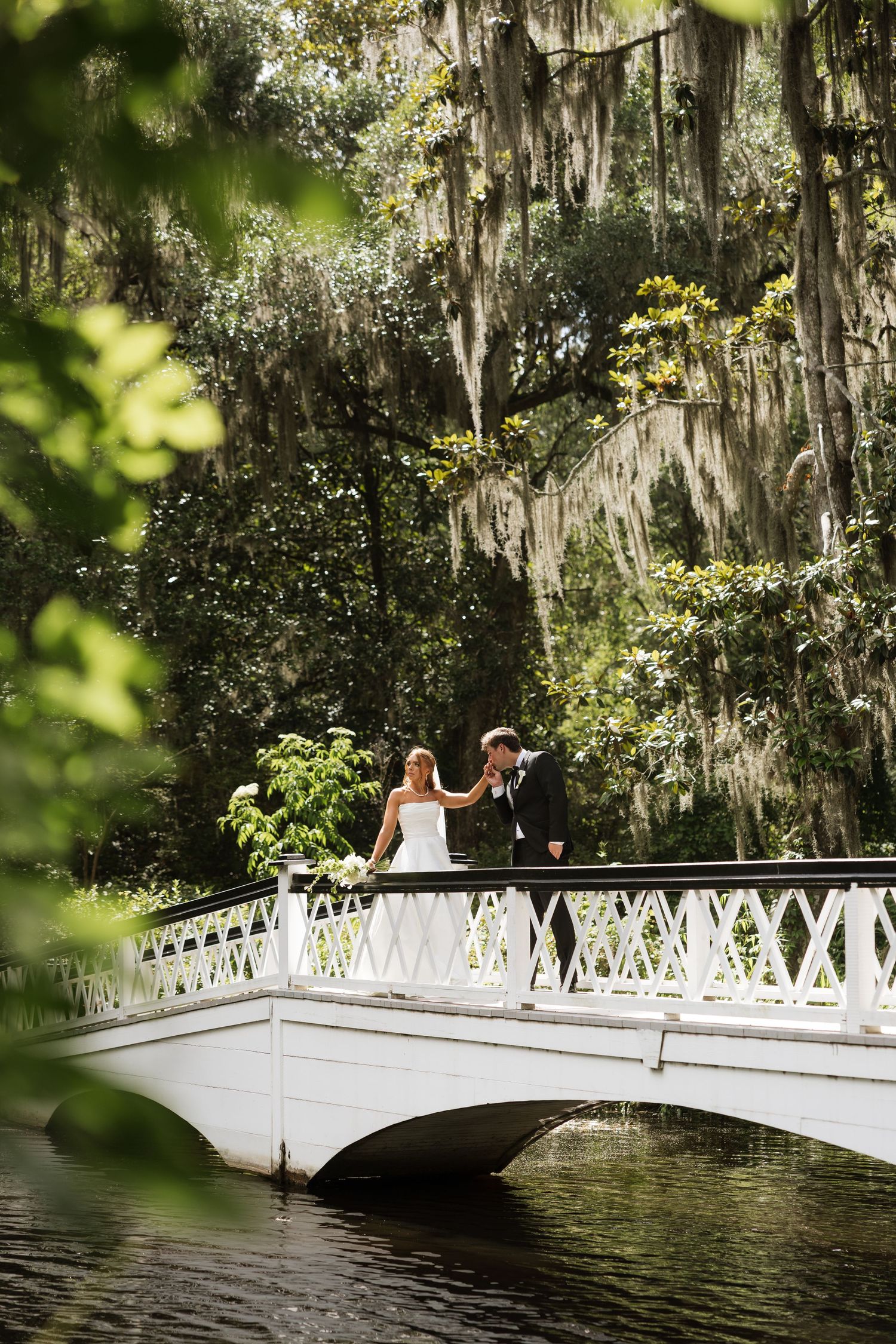 Bride and groom holding hands on a white bridge surrounded by lush trees draped with Spanish moss.