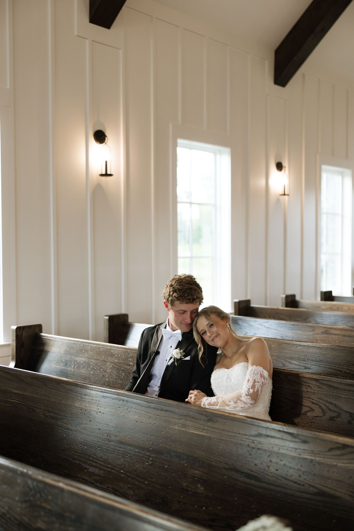 Bride and groom sitting together in wooden church pews, smiling at camera on wedding day.
