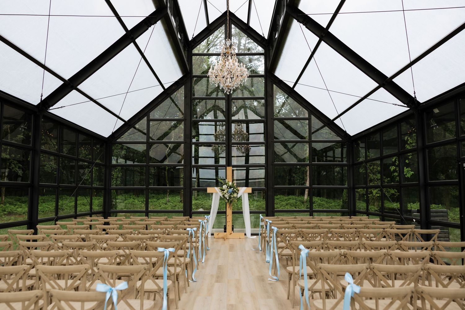 Elegant glass greenhouse wedding venue with chandelier, wooden cross-back chairs, and floral altar decor.