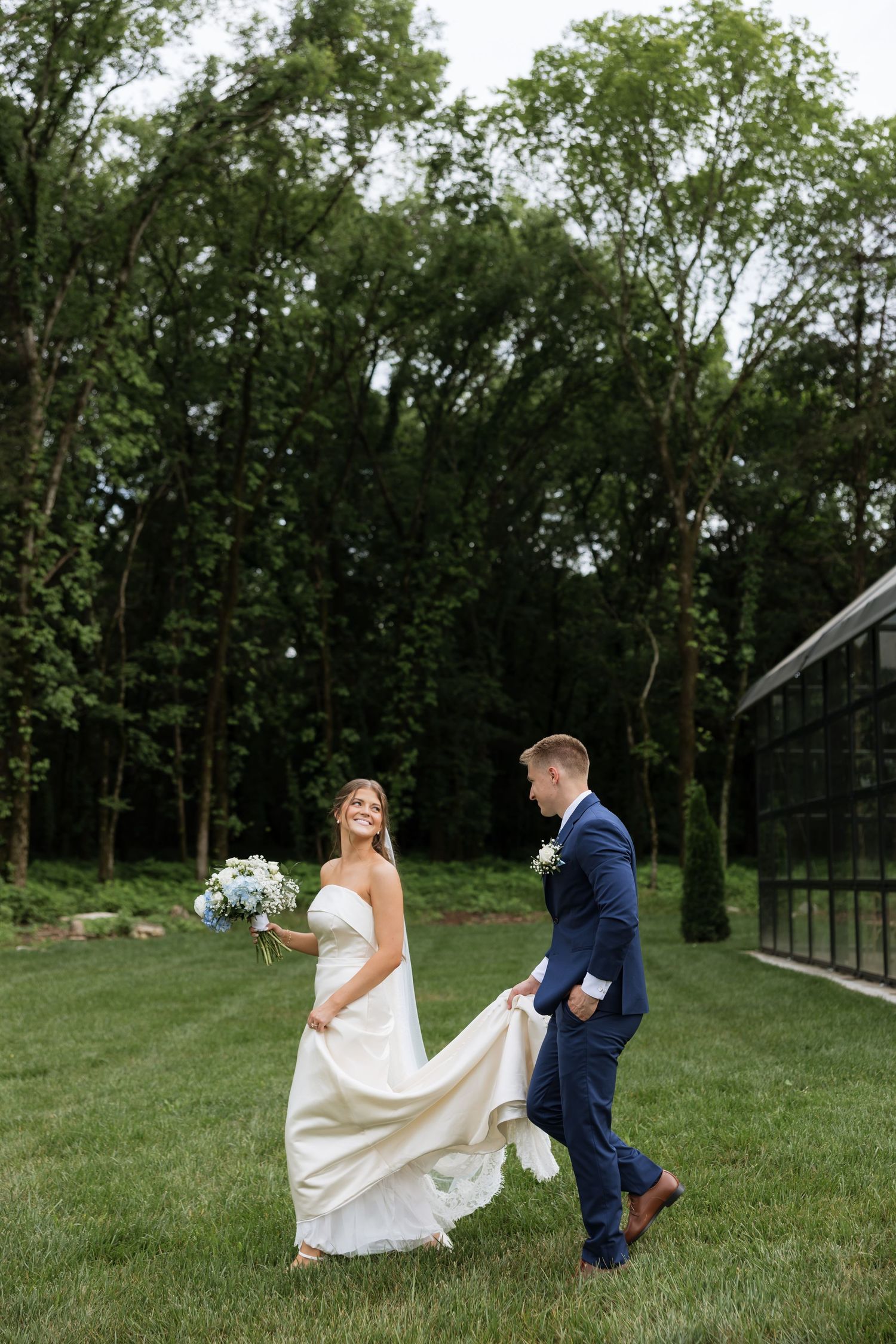 Bride and groom walk on green lawn, groom carrying wedding dress train, bride holding blue bouquet.