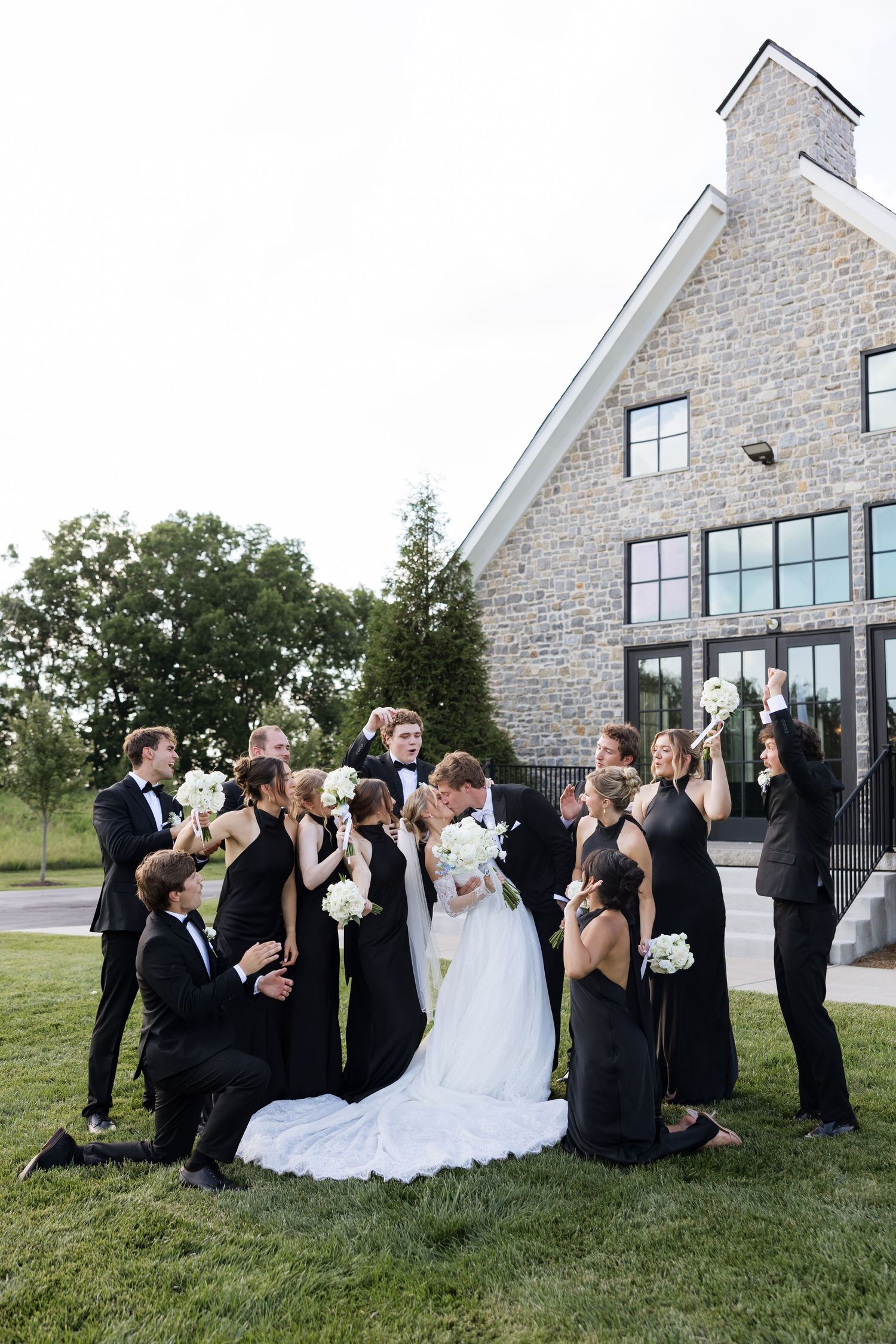 Wedding party in black attire surrounds kissing bride and groom outside elegant stone building with white flowers.