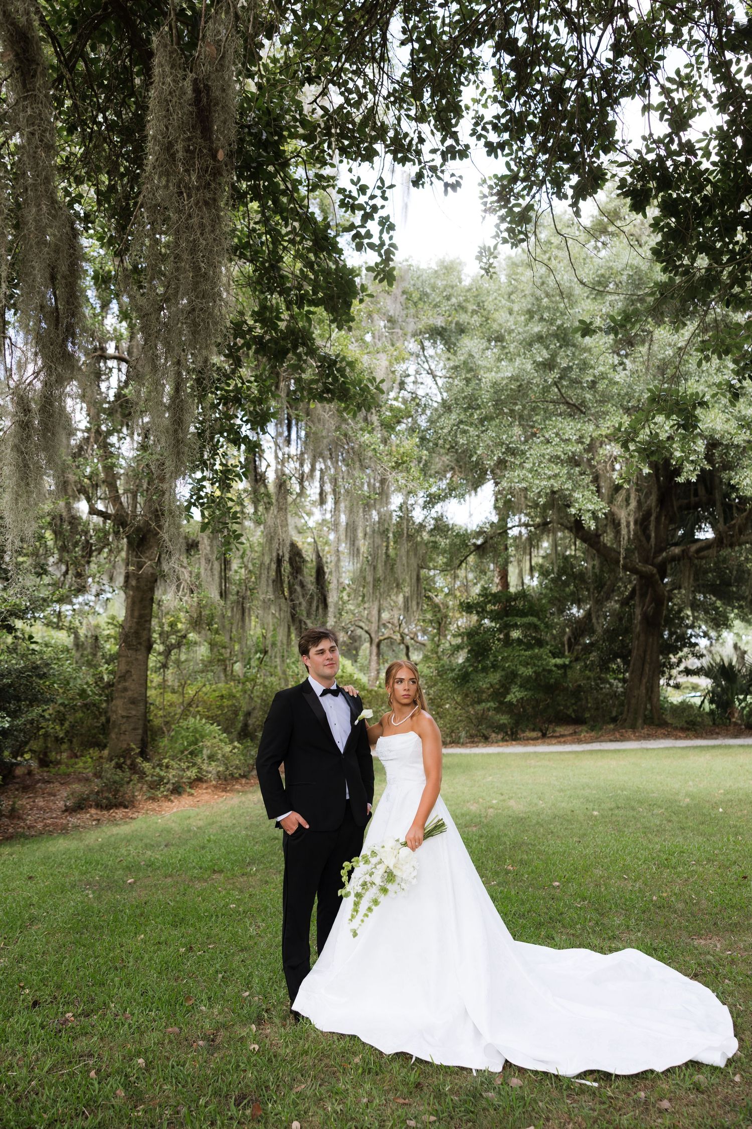 Bride and groom pose under Spanish moss-draped oak trees on lush green lawn in classic wedding attire.