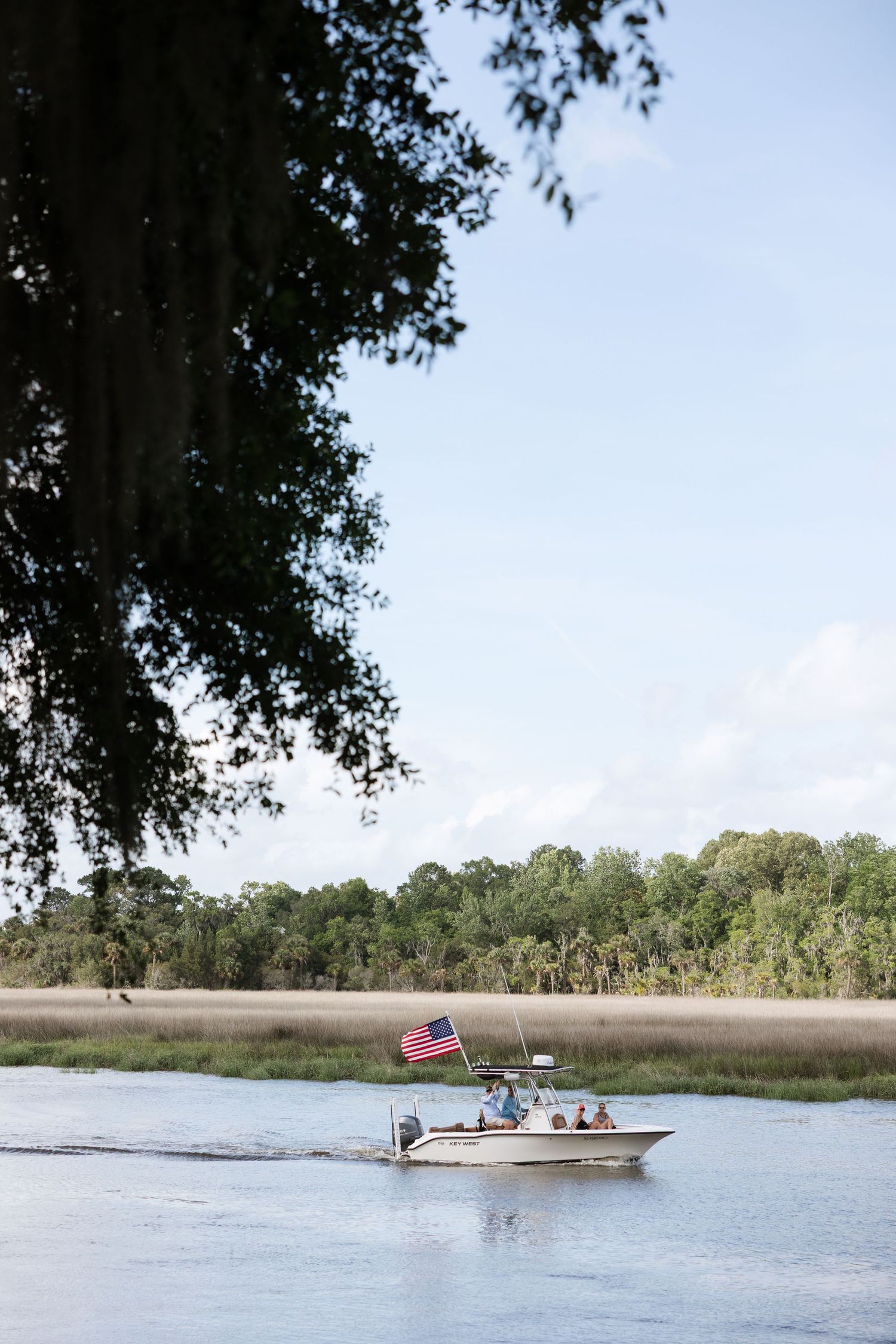 A motorboat with an American flag cruises through a scenic Southern marsh waterway framed by oak trees.