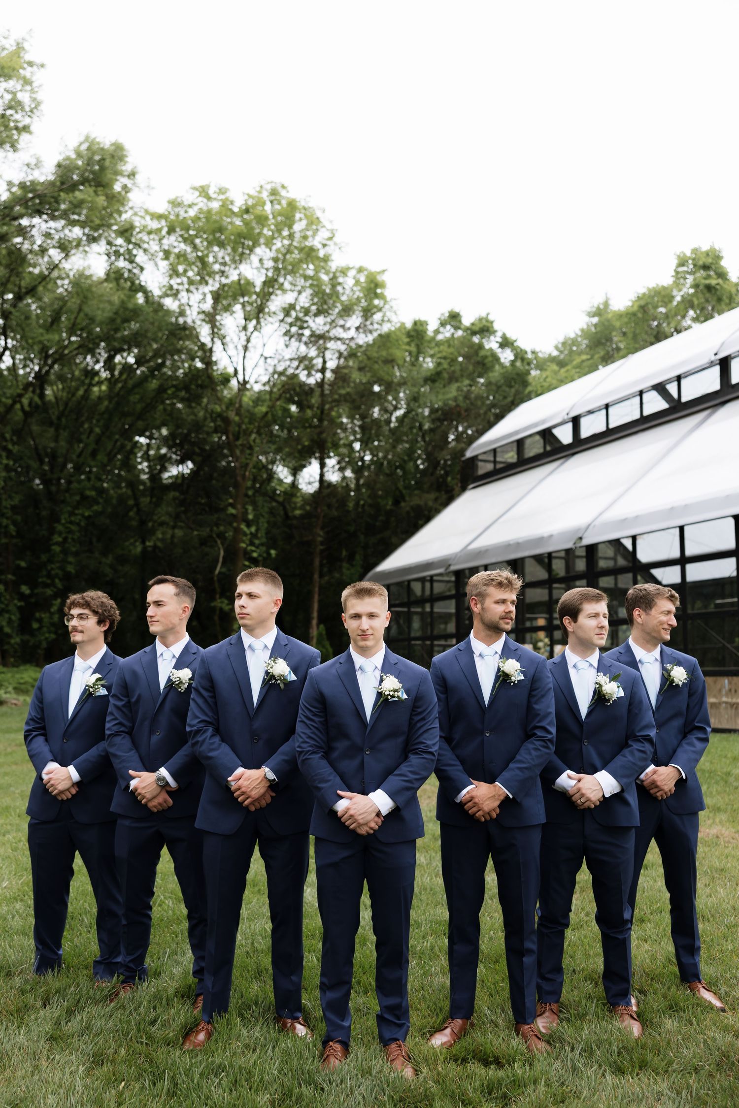 Six groomsmen in navy blue suits with boutonnieres standing outdoors at wedding venue.