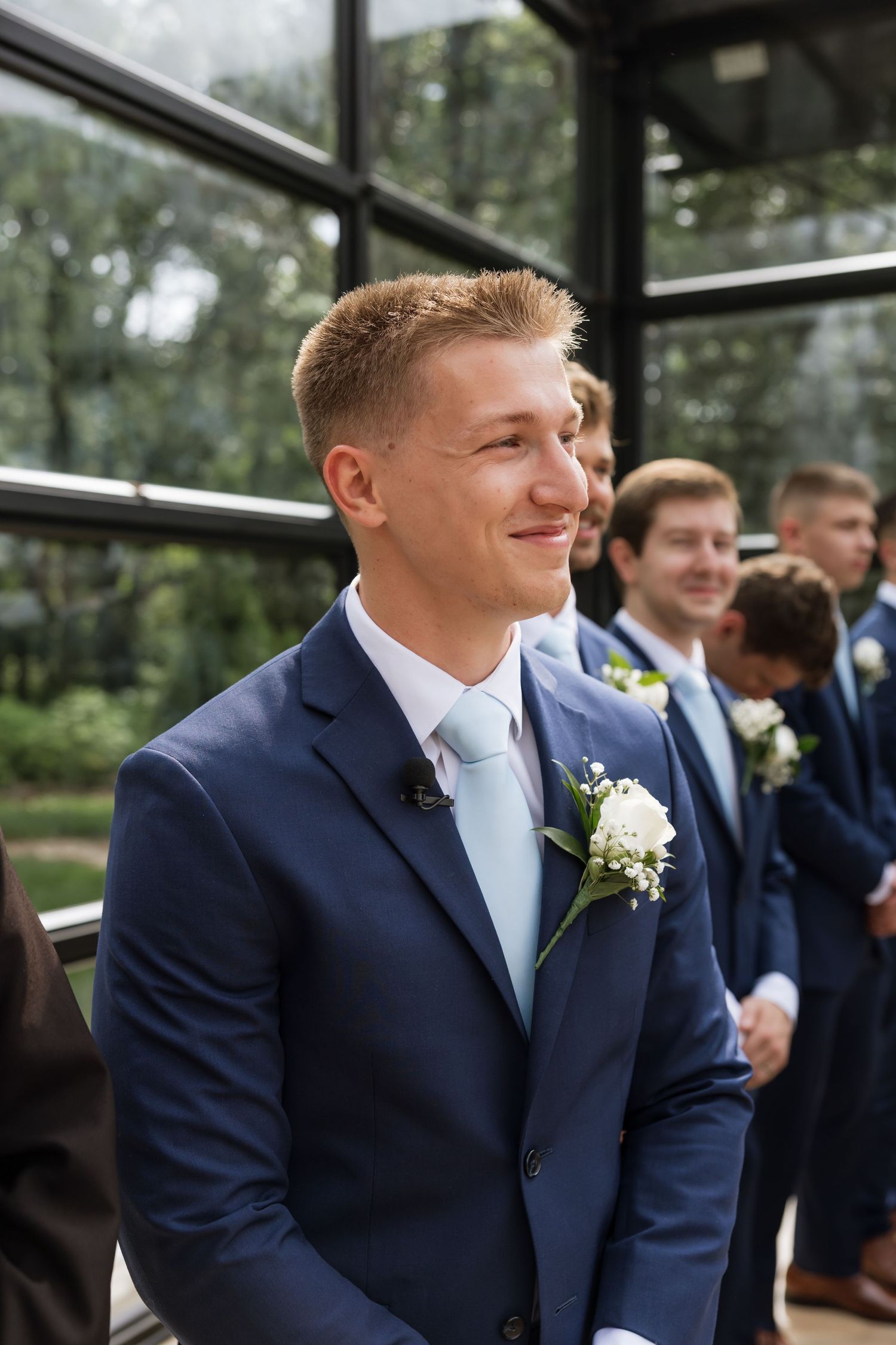 Smiling groom in navy blue suit with white boutonniere stands with groomsmen during wedding ceremony.
