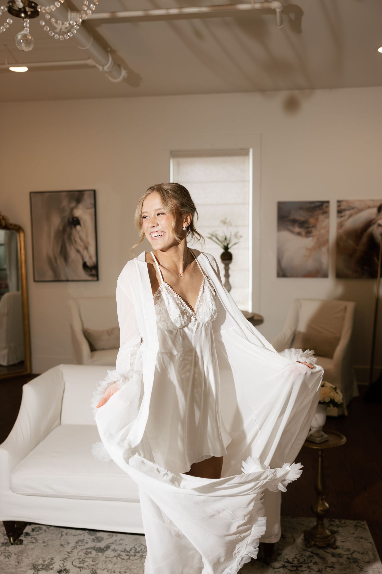 Joyful bride twirling in white bridal robe inside elegant room with horse artwork.