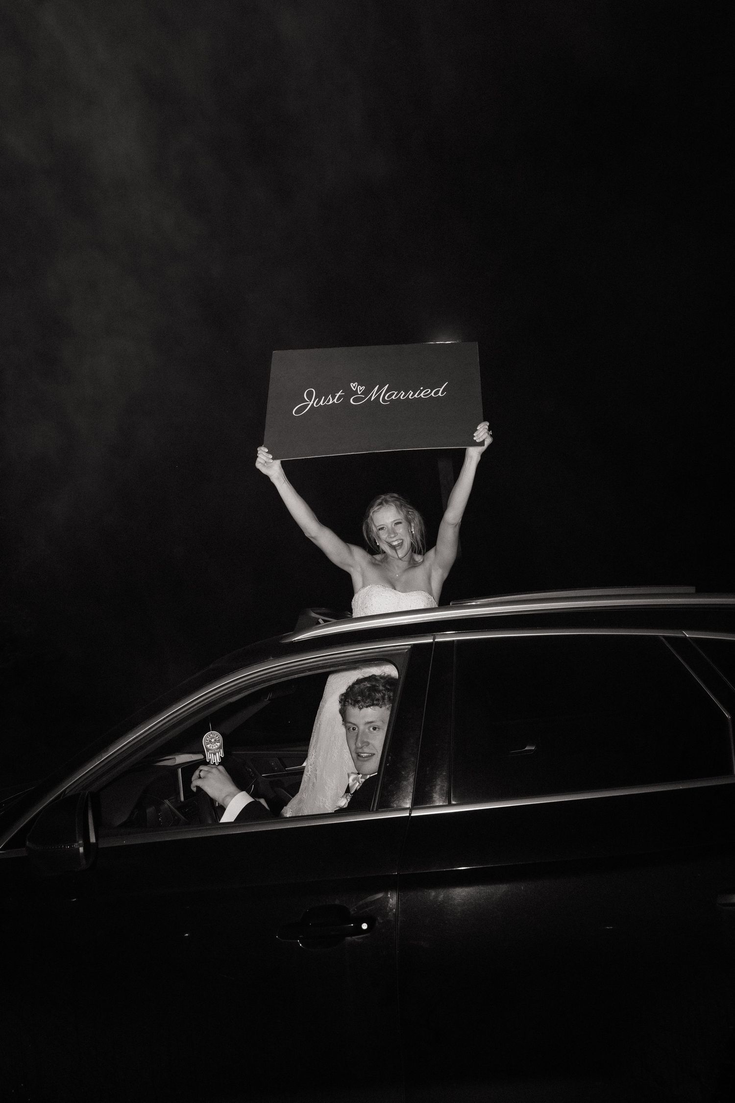Newlyweds celebrating in car with Just Married sign raised through sunroof at night in black and white.