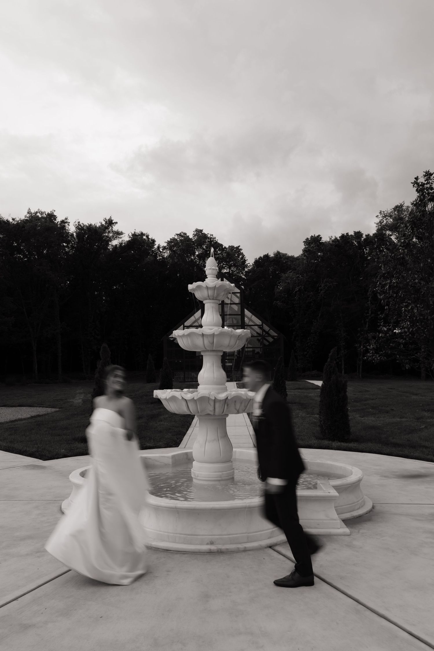 Bride and groom walking past elegant white tiered fountain in black and white motion-blur wedding photo.