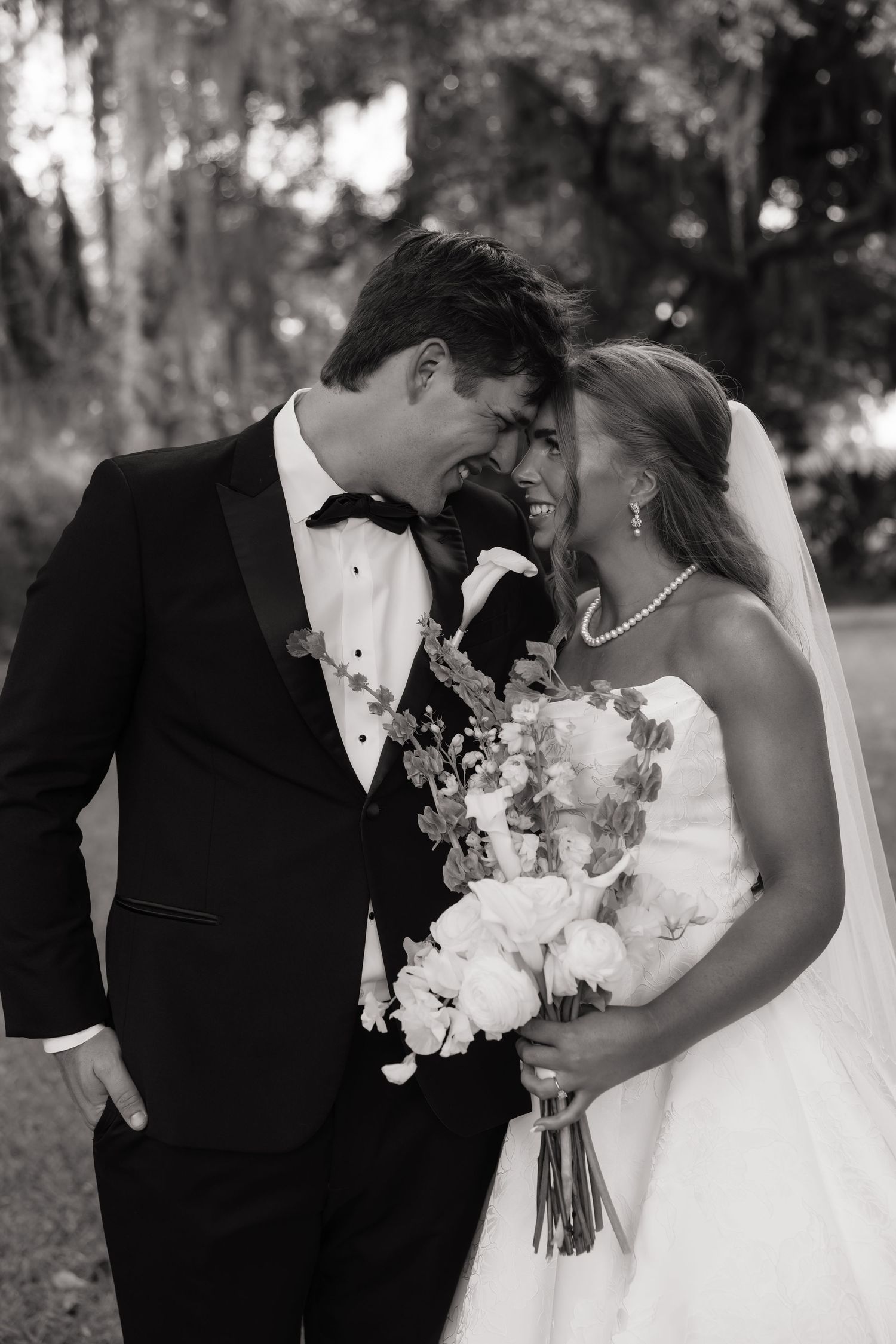 Black and white photo of smiling bride and groom touching foreheads, bride holding floral bouquet outdoors.
