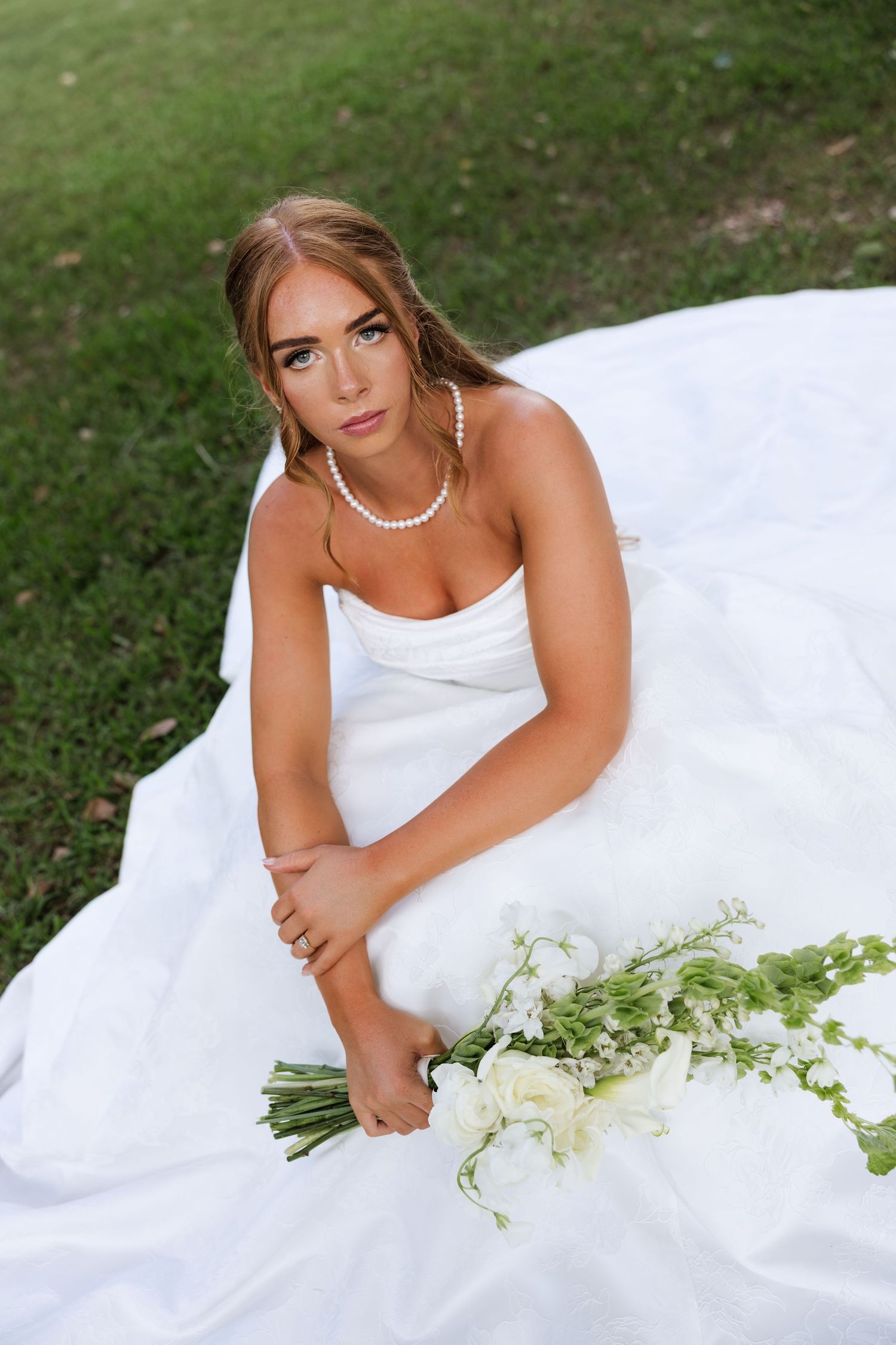 Bride in white strapless gown sitting on green grass, wearing pearl necklace, holding white floral bouquet.