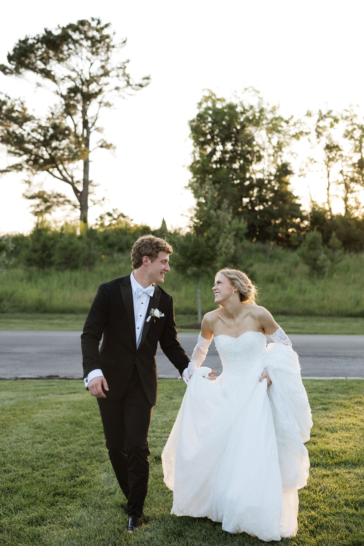 Bride and groom walk hand-in-hand on green lawn at golden hour, smiling at each other in wedding attire.