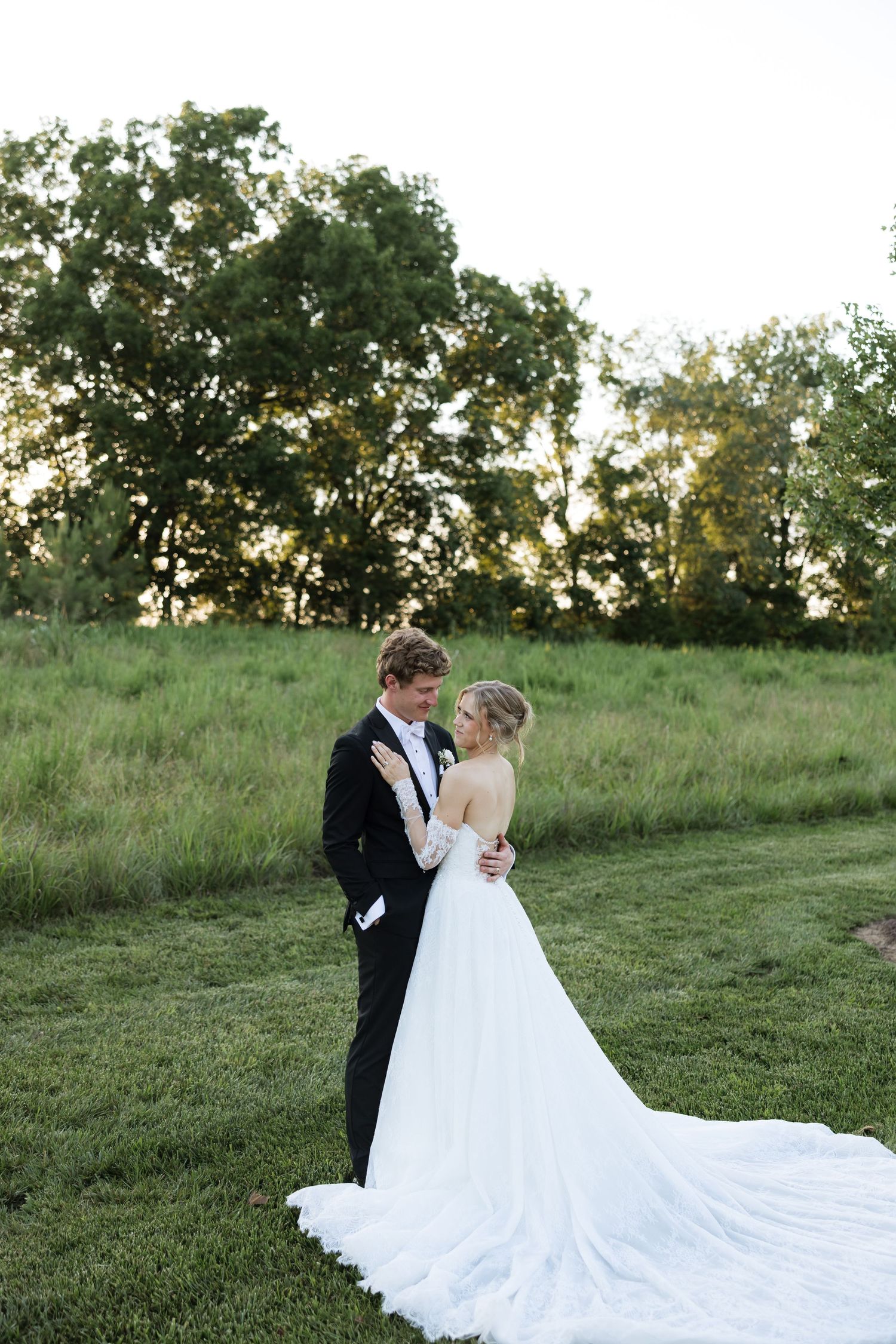 Bride and groom embrace at sunset in lush green field, bride wearing elegant white gown with long train.