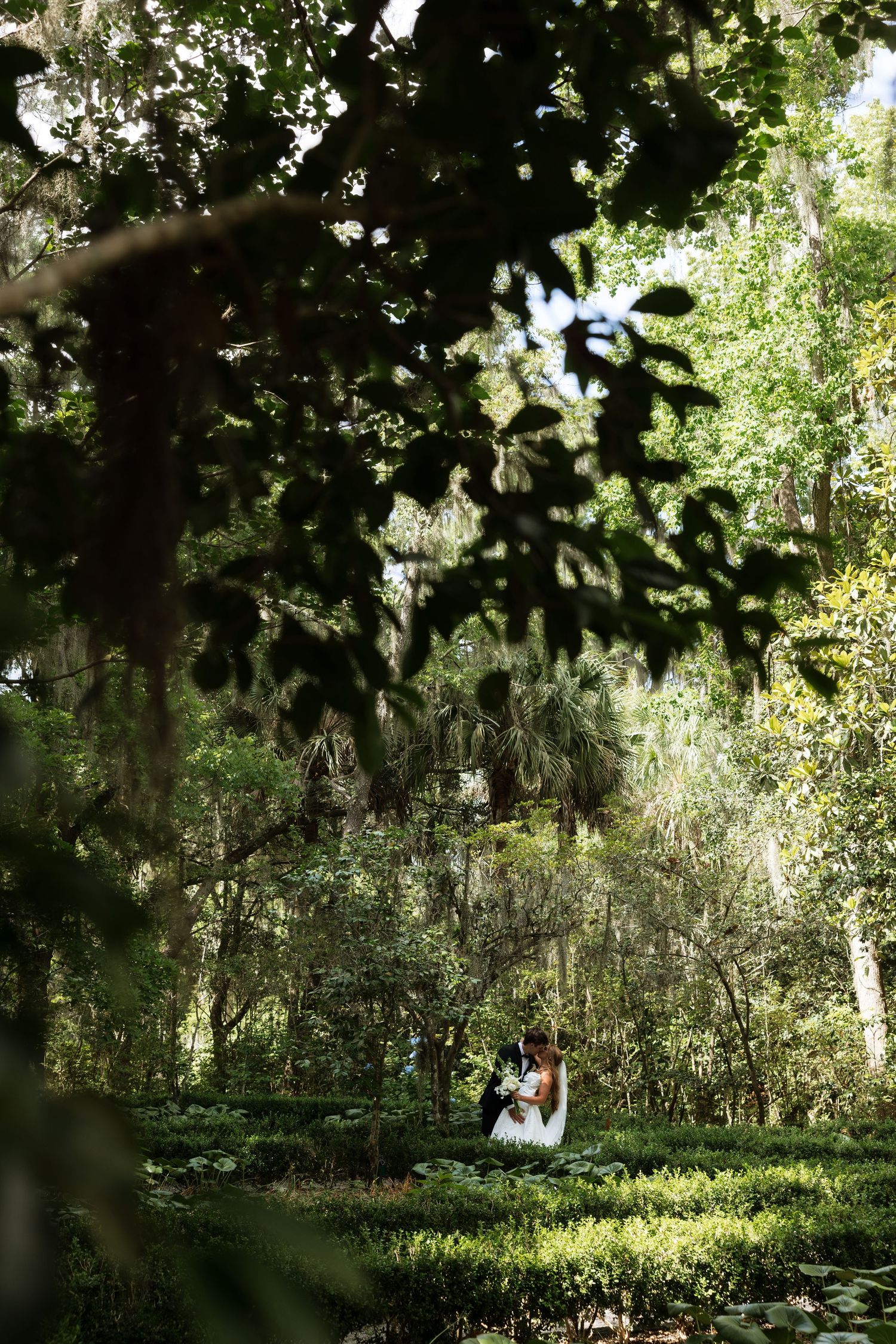 Bride and groom embrace in a lush green garden surrounded by moss-draped trees.