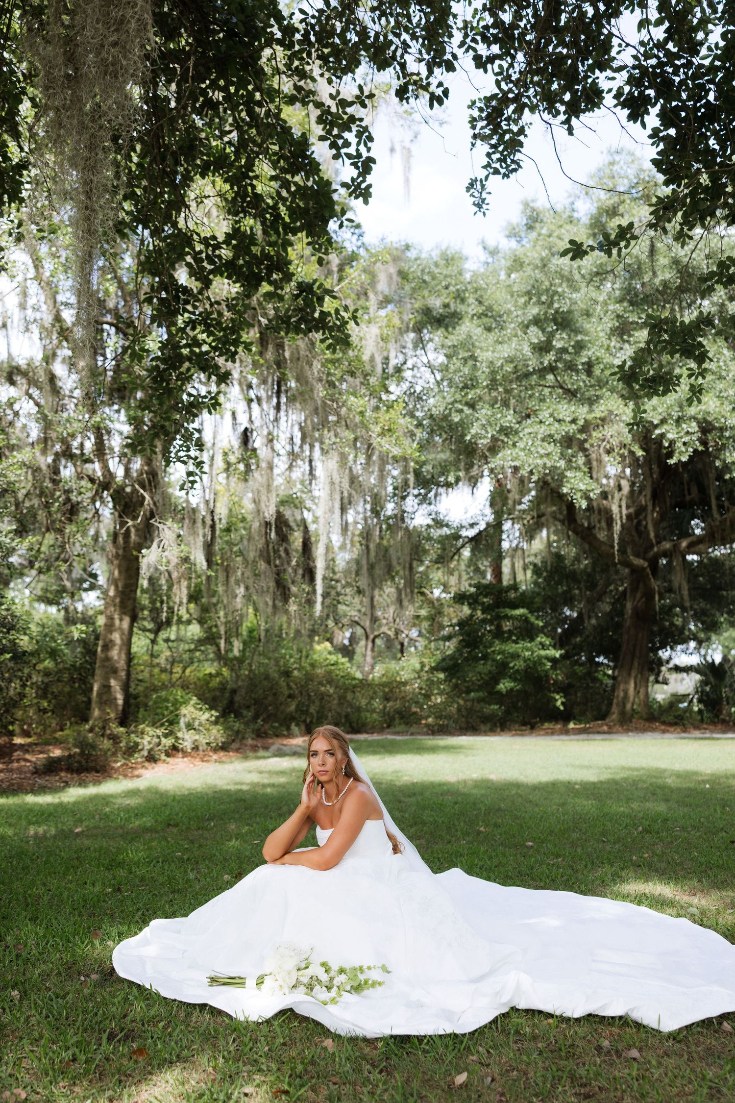 Bride in a flowing white gown sits on lush green grass beneath Spanish moss-draped trees holding a white bouquet.