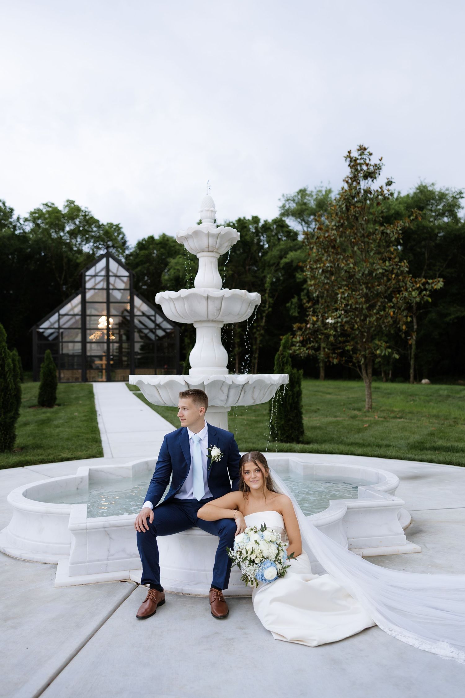 Bride and groom pose elegantly by white decorative fountain at outdoor wedding venue at dusk.