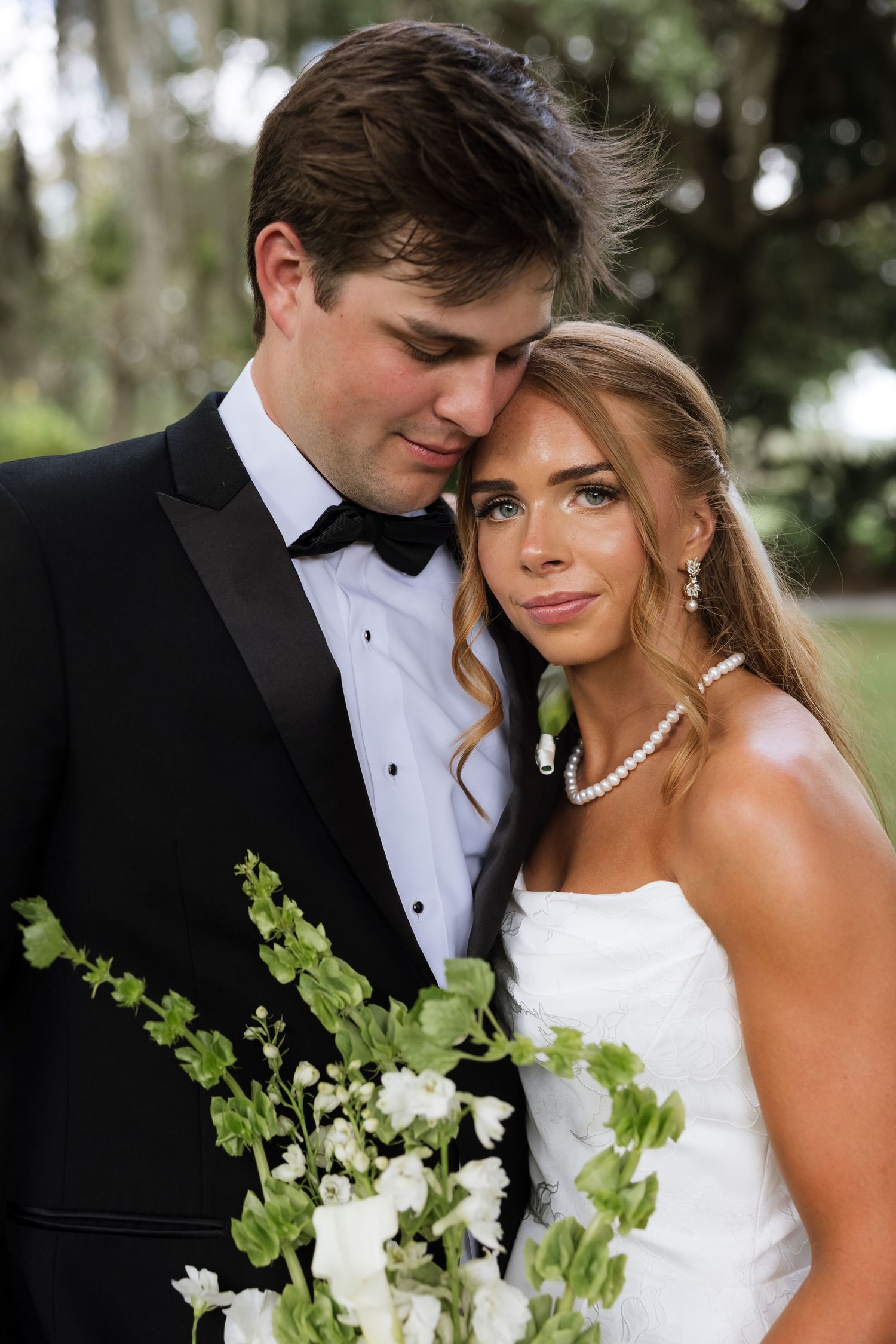 Bride and groom share an intimate moment, she in a strapless gown with pearls, he in a black tuxedo, holding white floral bouquet.
