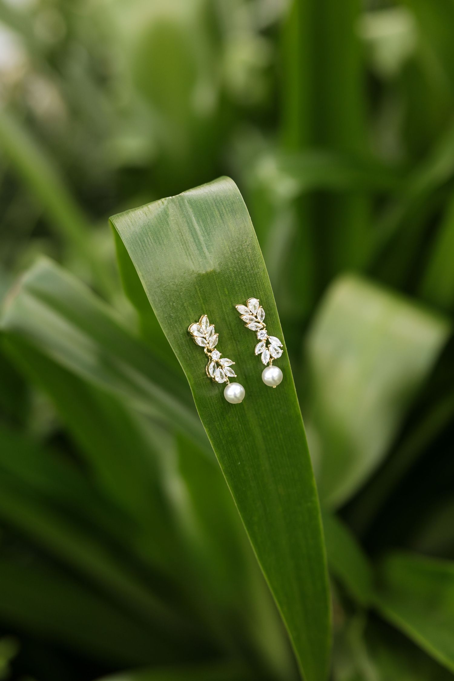 Gold leaf pearl drop earrings displayed on a vibrant green tropical leaf in a lush garden setting.