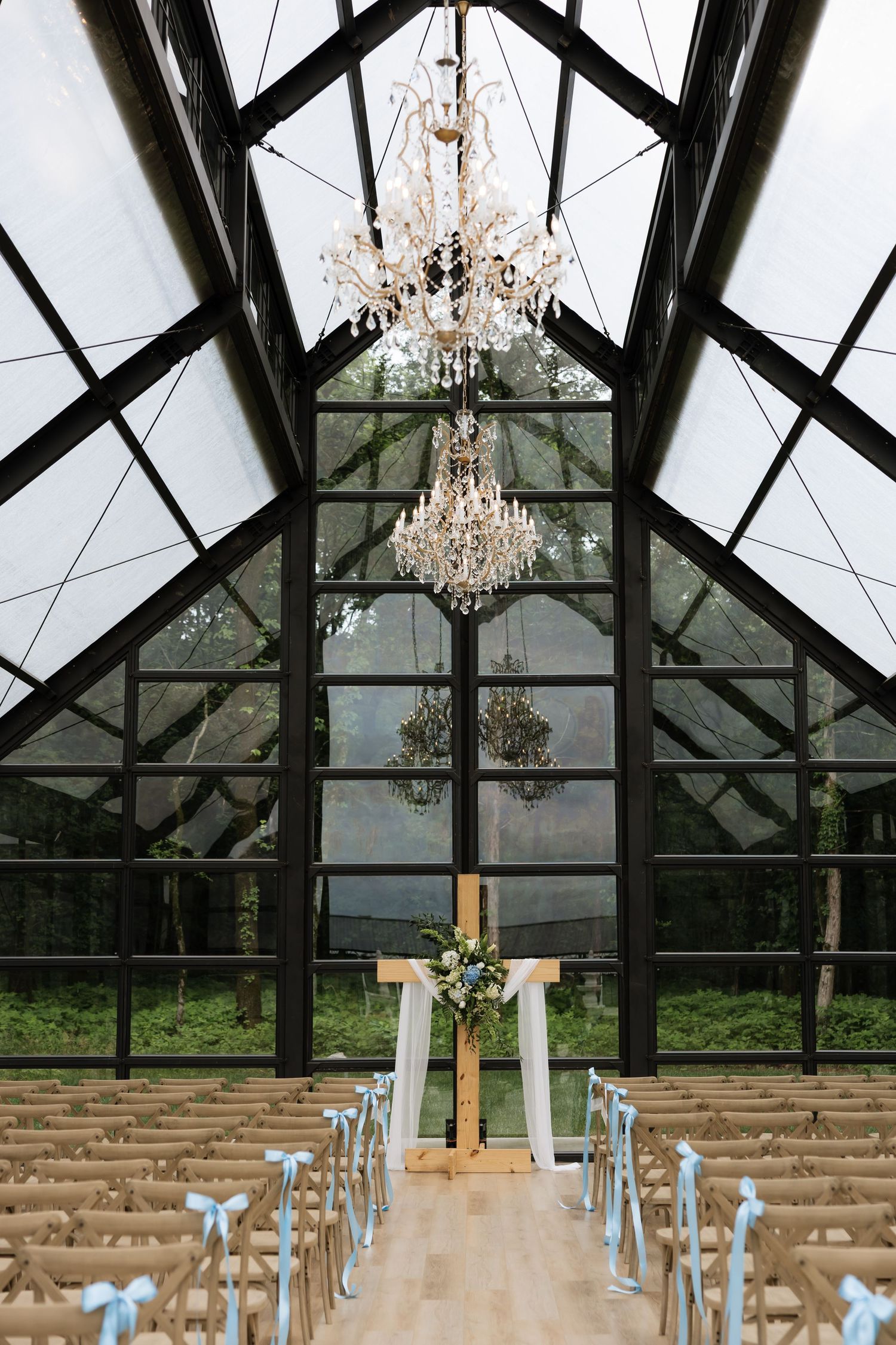 Elegant greenhouse wedding venue with crystal chandeliers, wooden arch, and rows of chairs with blue ribbon bows.