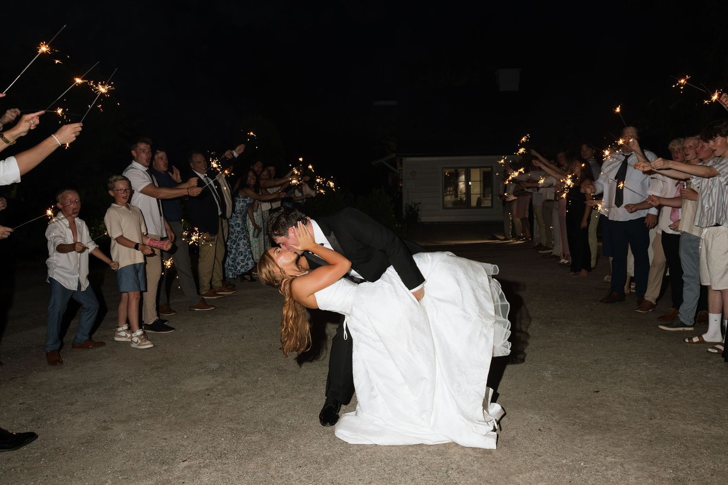 Bride and groom share a romantic dip kiss during sparkler exit at outdoor wedding reception at night.