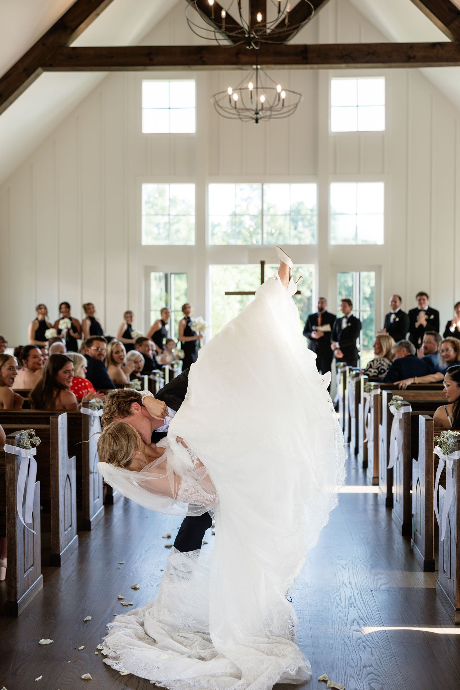 Groom dips bride in white gown during wedding ceremony kiss in elegant white chapel with wooden pews and chandelier.
