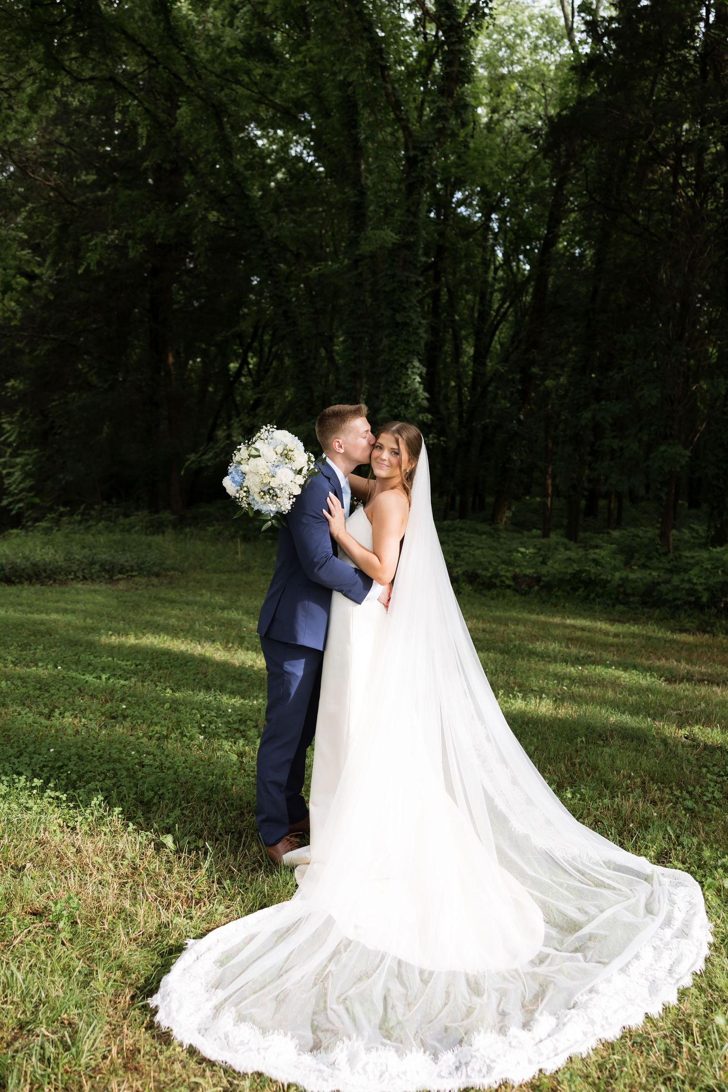 Bride and groom embrace in lush green outdoor setting, bride wearing elegant white gown with long veil and bouquet.