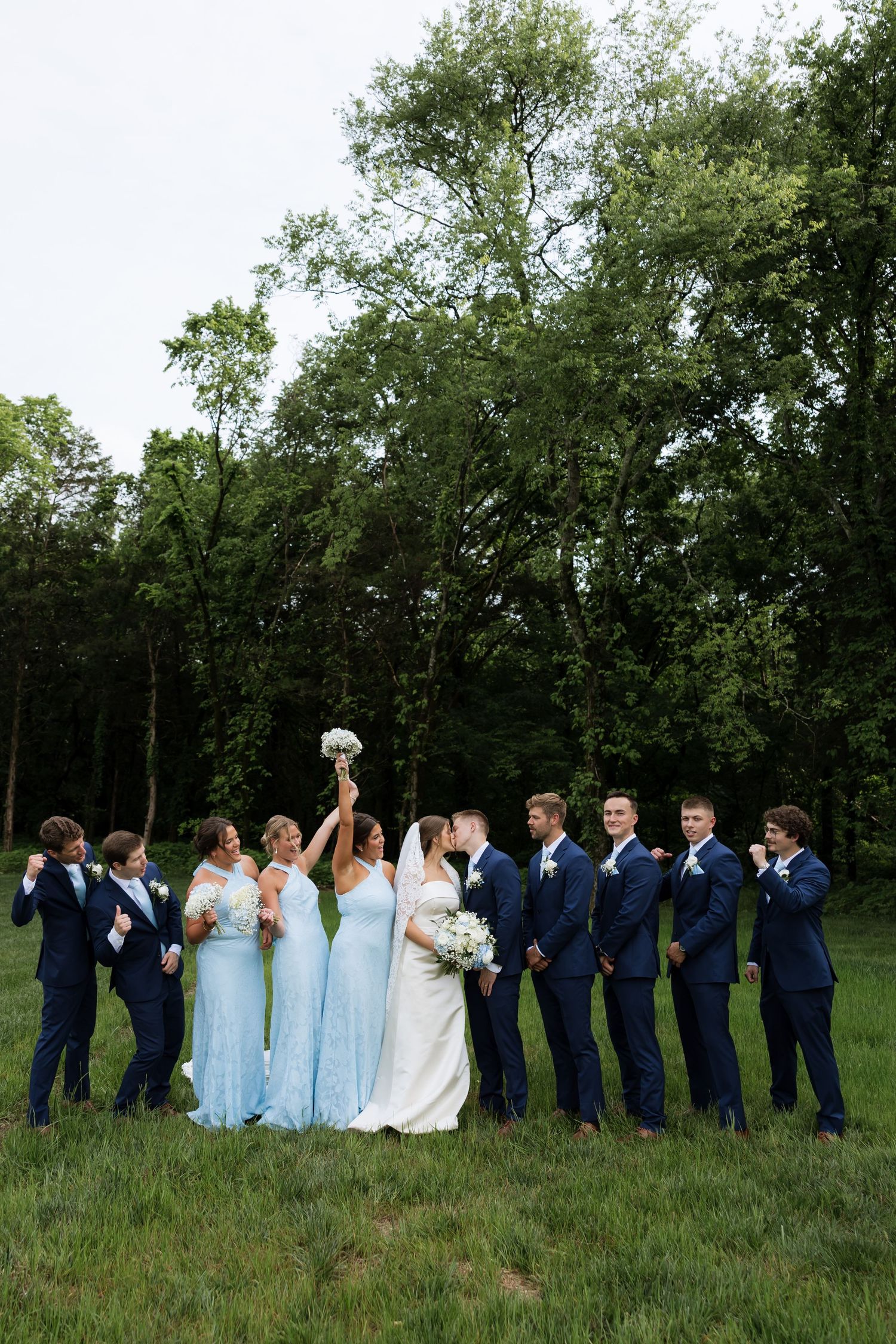 Wedding party poses outdoors, bride and groom kissing, bridesmaids in light blue gowns, groomsmen in navy suits.