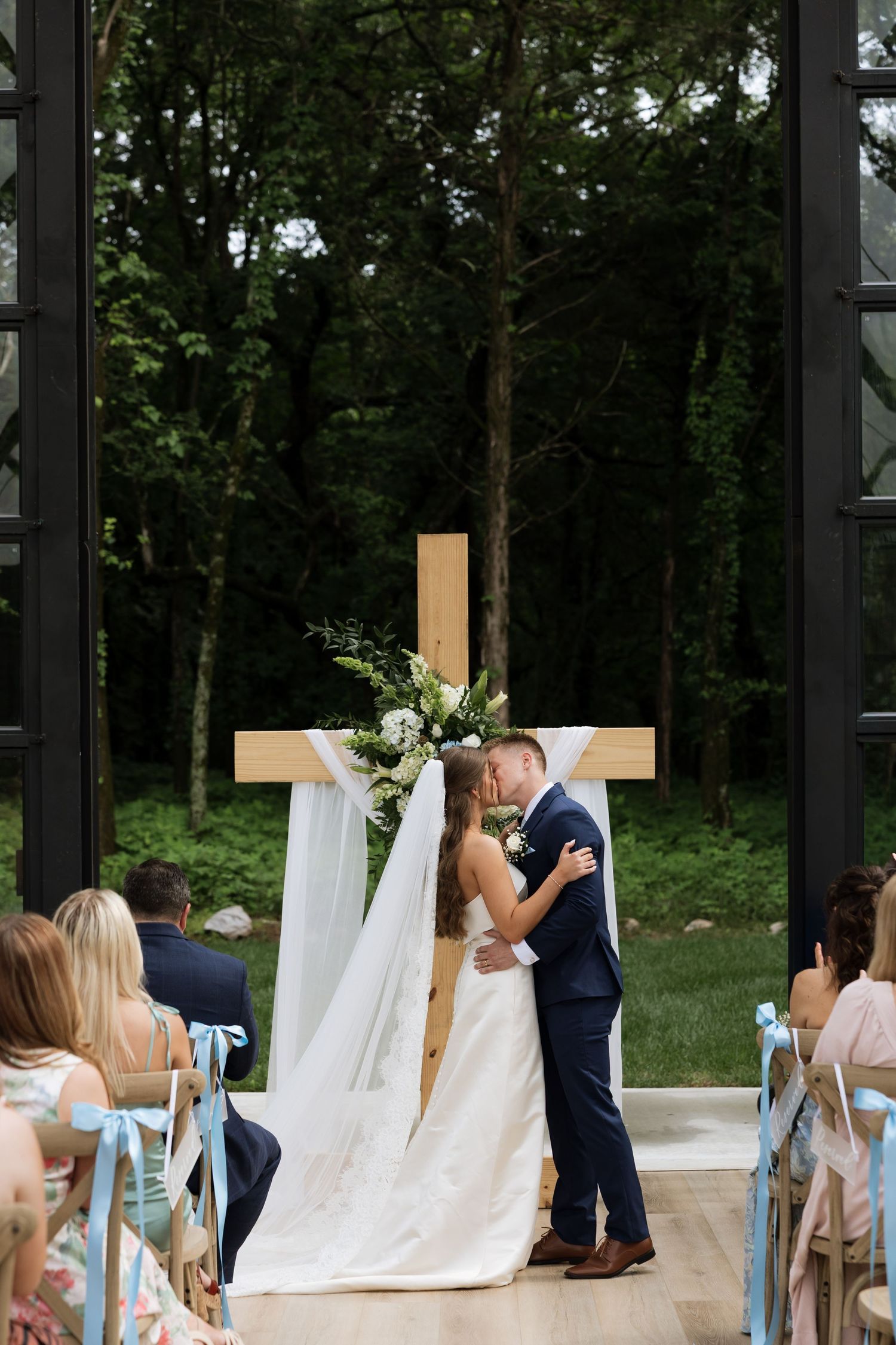 Newlyweds share first kiss at altar with wooden cross and floral decor in a glass chapel overlooking lush forest.