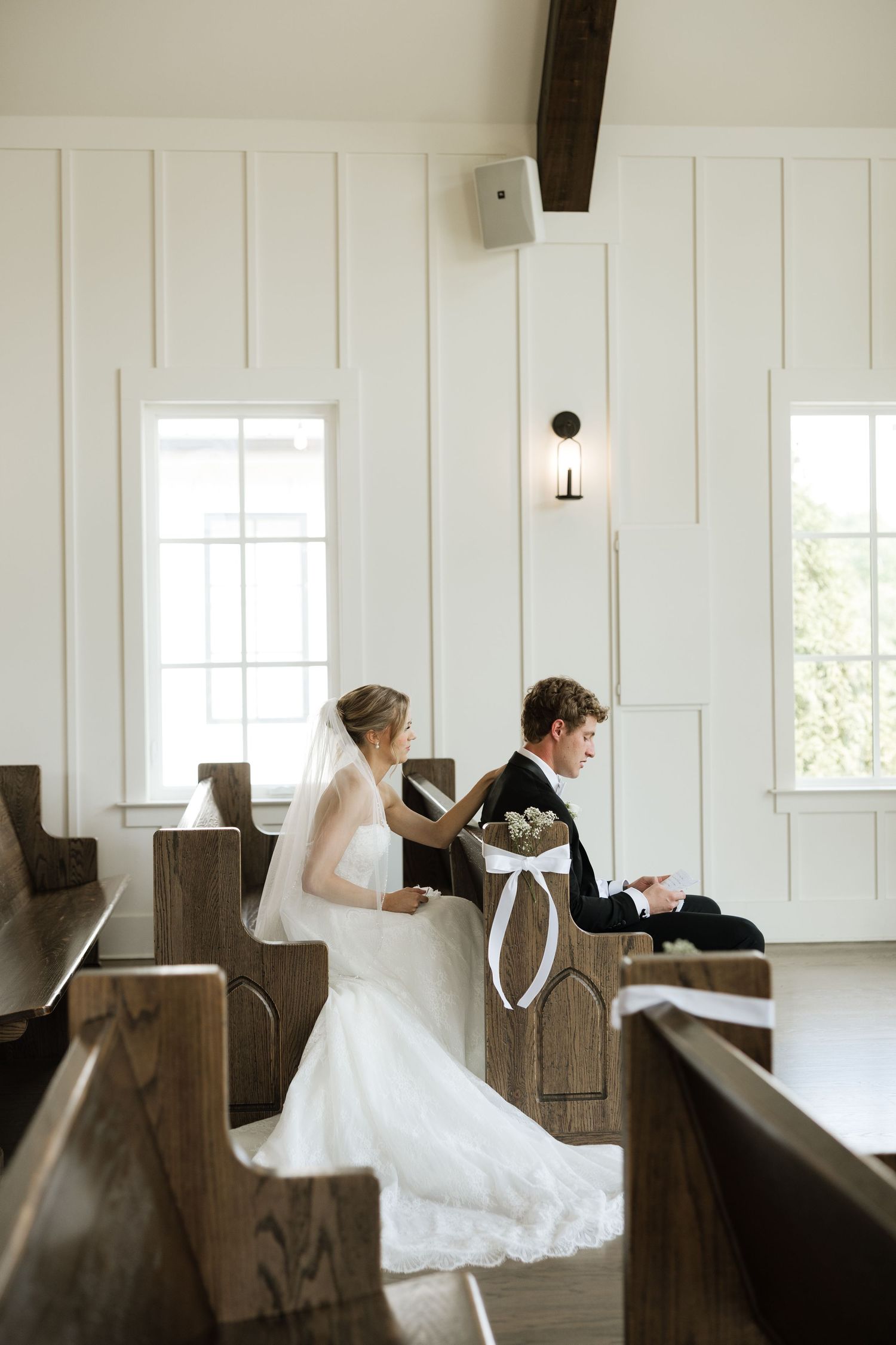Bride and groom sit together in white chapel pews during intimate wedding ceremony moment.