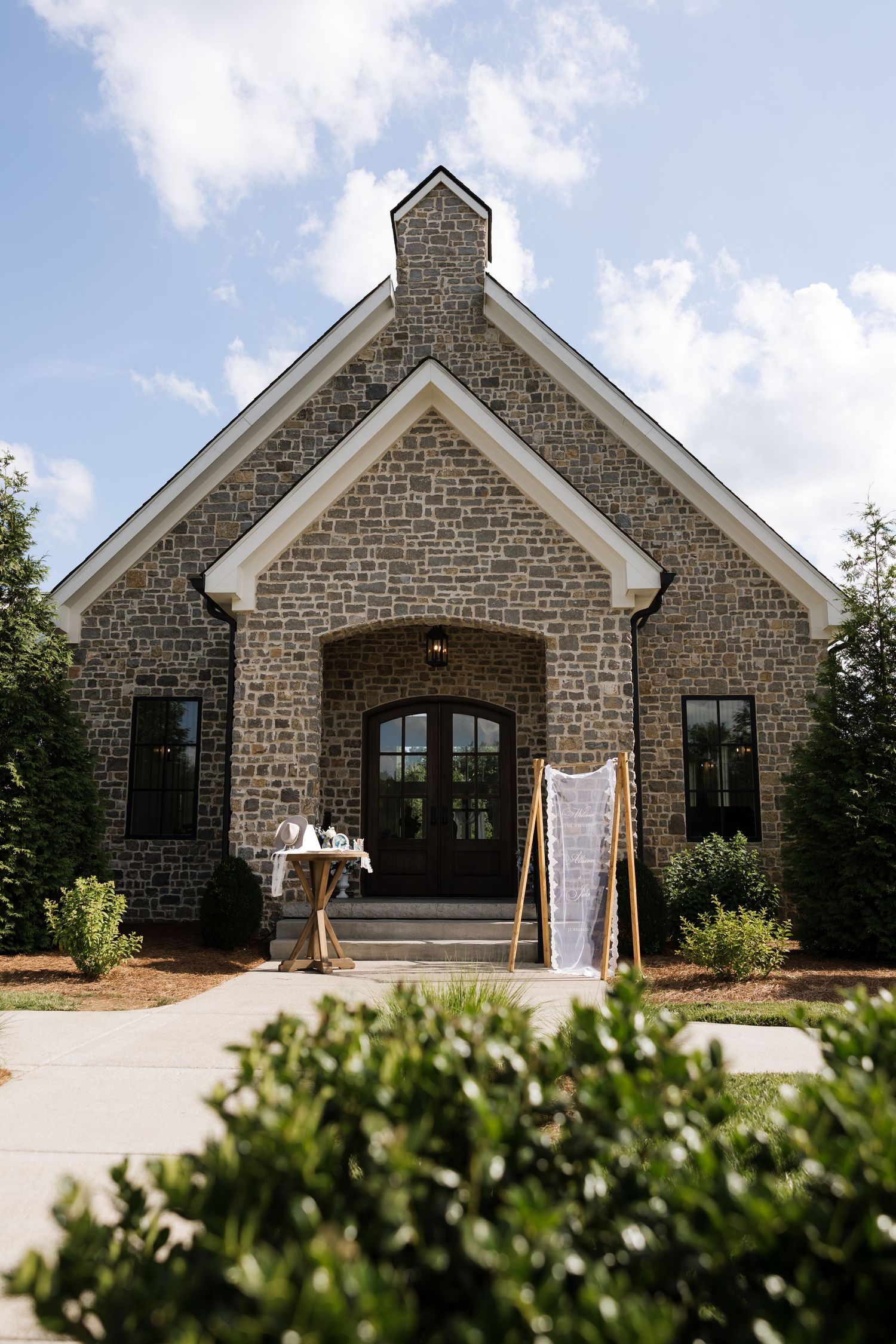 Stone chapel wedding venue with arched entrance, French doors, and elegant signage surrounded by lush greenery under blue skies.