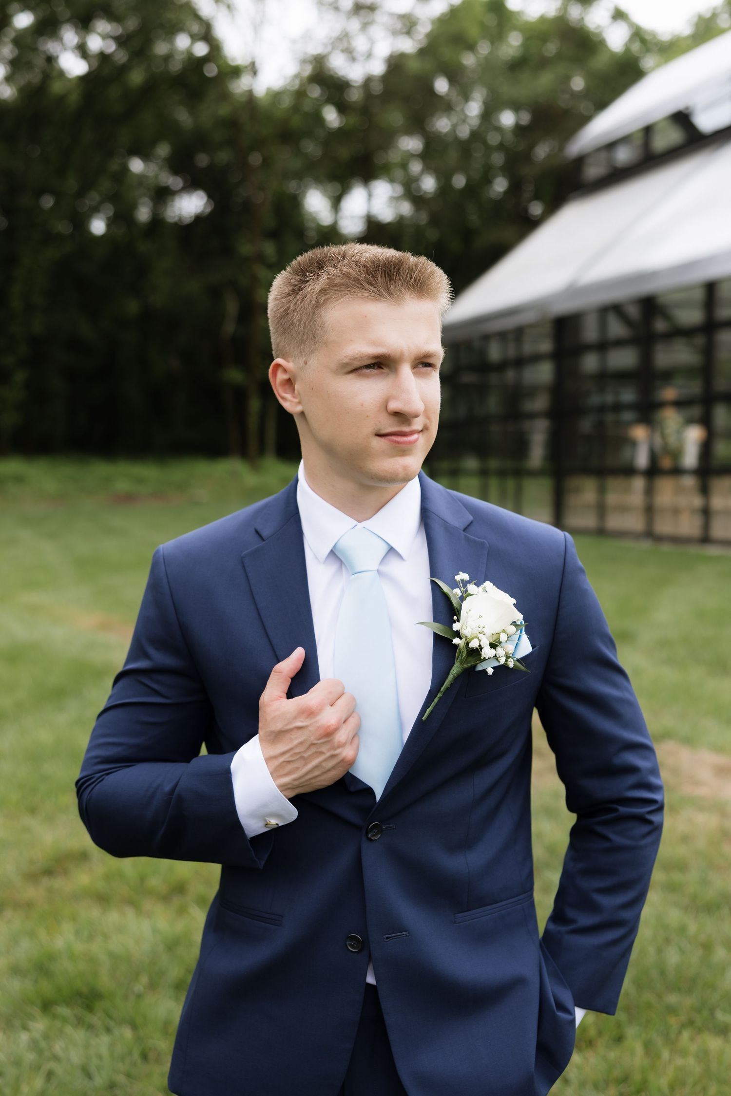 Groom in navy blue suit with white boutonniere and light blue tie poses outdoors at wedding venue.