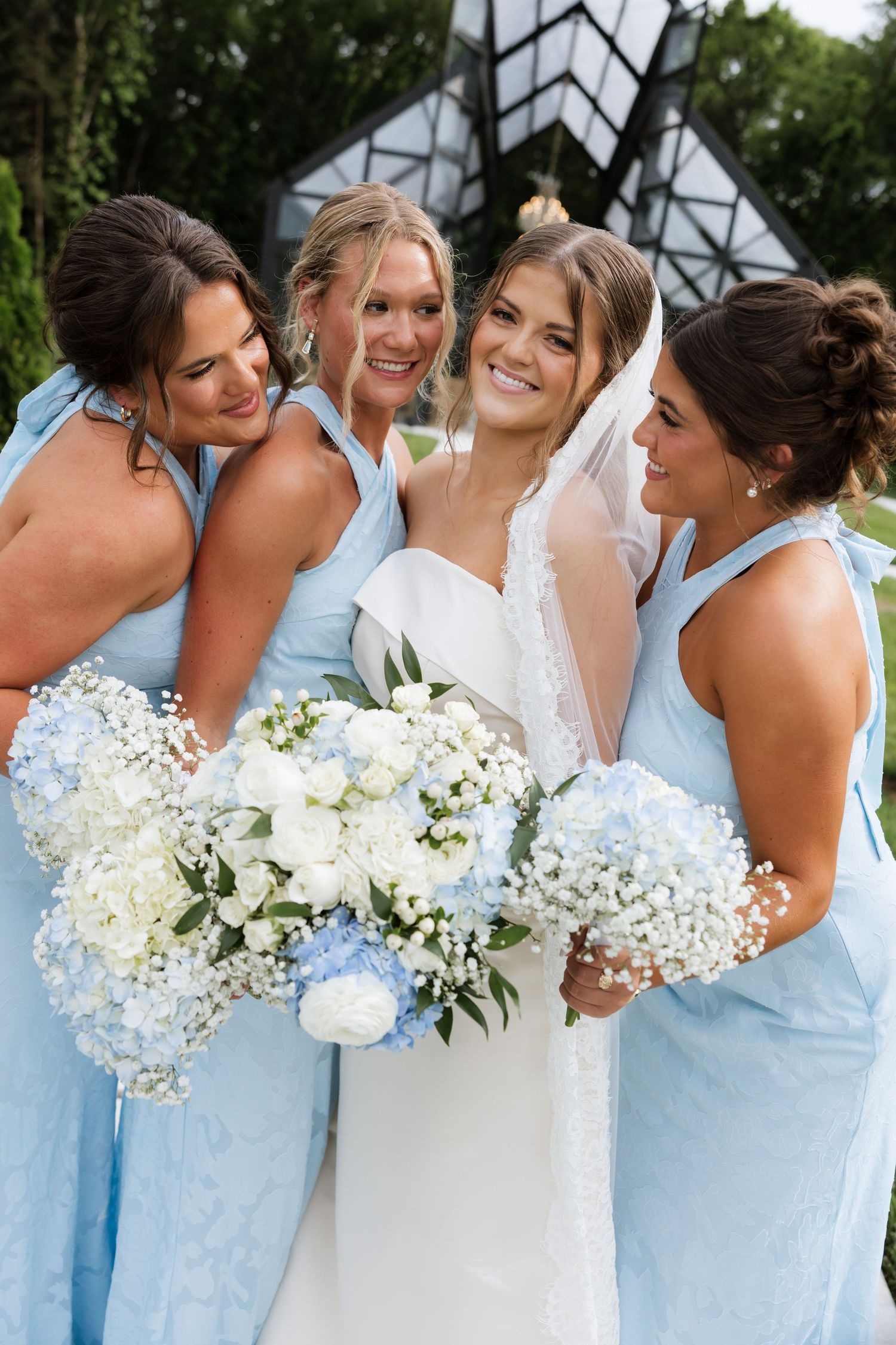 Bride in white dress and veil surrounded by smiling bridesmaids in blue holding white and blue bouquets.