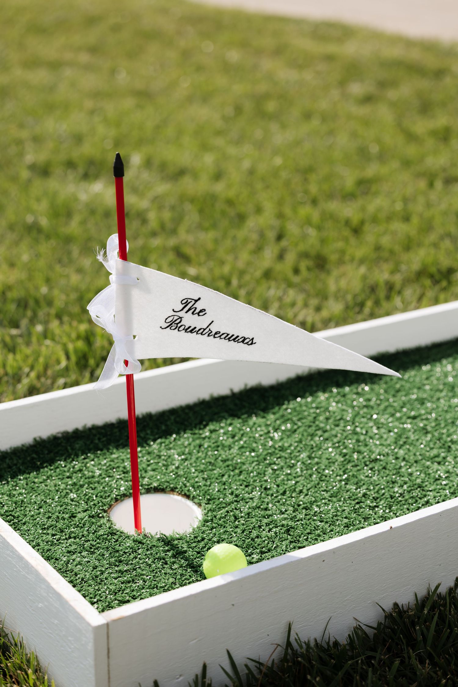Custom mini golf hole with white wooden border, green turf, and personalized pennant flag reading The Boudreauxs.