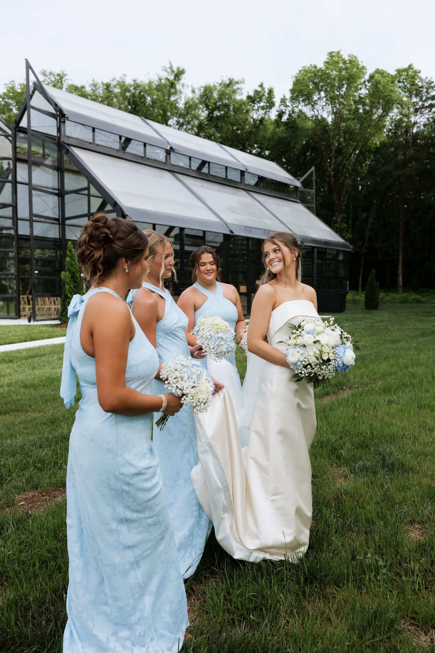 Bride in white gown with bridesmaids in light blue dresses holding bouquets outside a glass greenhouse.