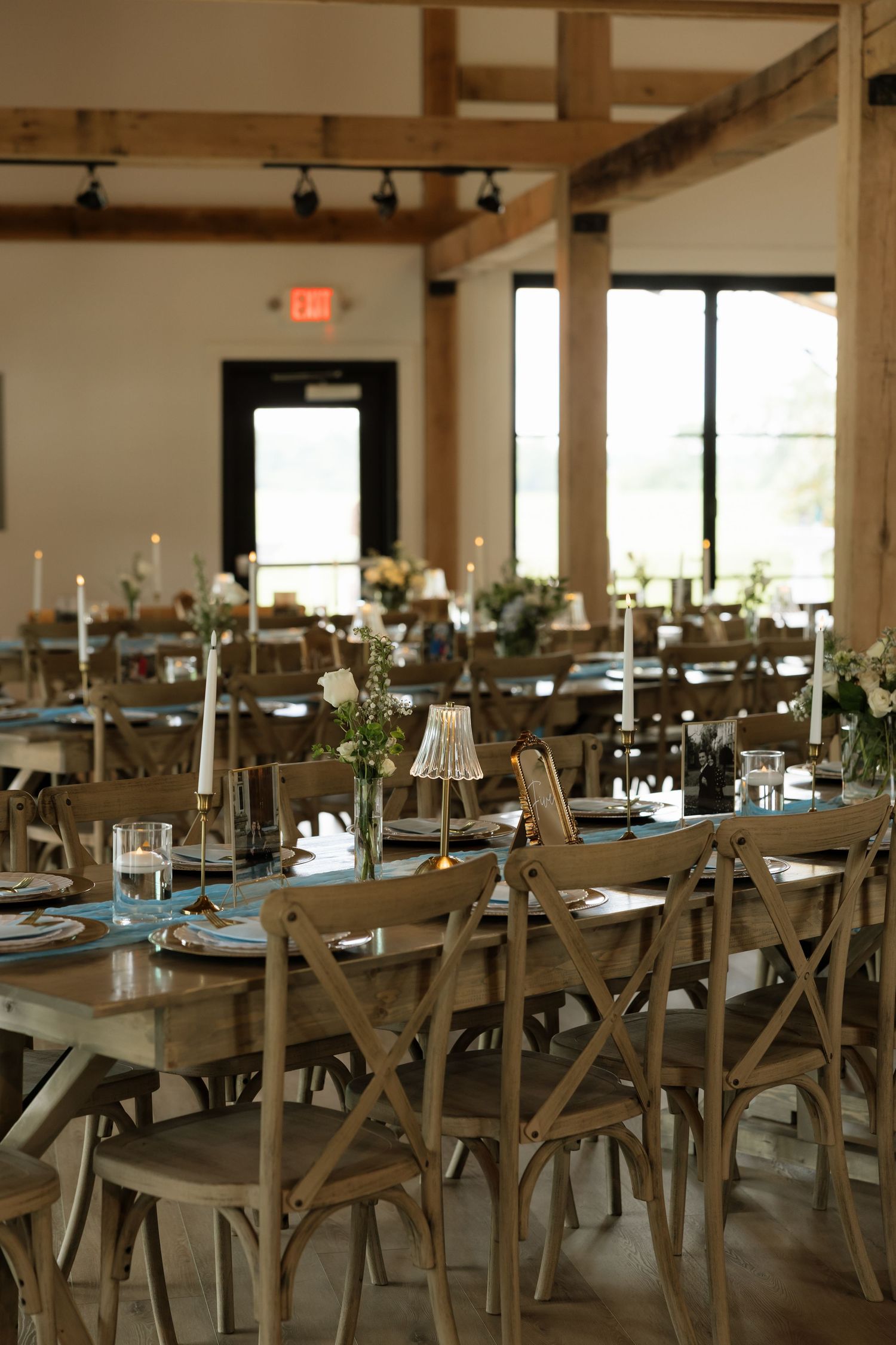 Rustic wedding reception hall with wooden farm tables, cross-back chairs, candles, and floral centerpieces in warm lighting.