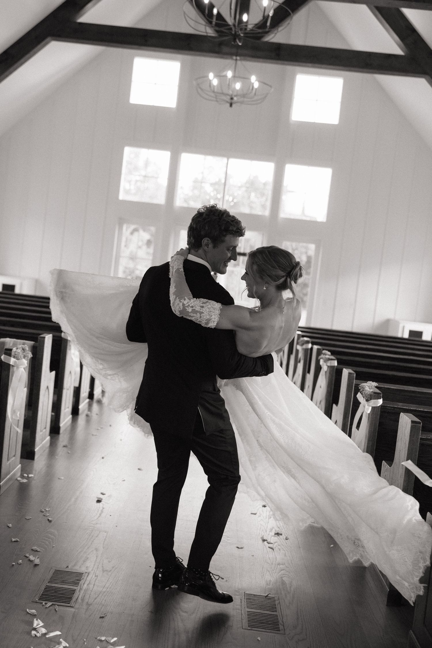 Black and white photo of groom lifting bride in white gown inside charming chapel aisle.