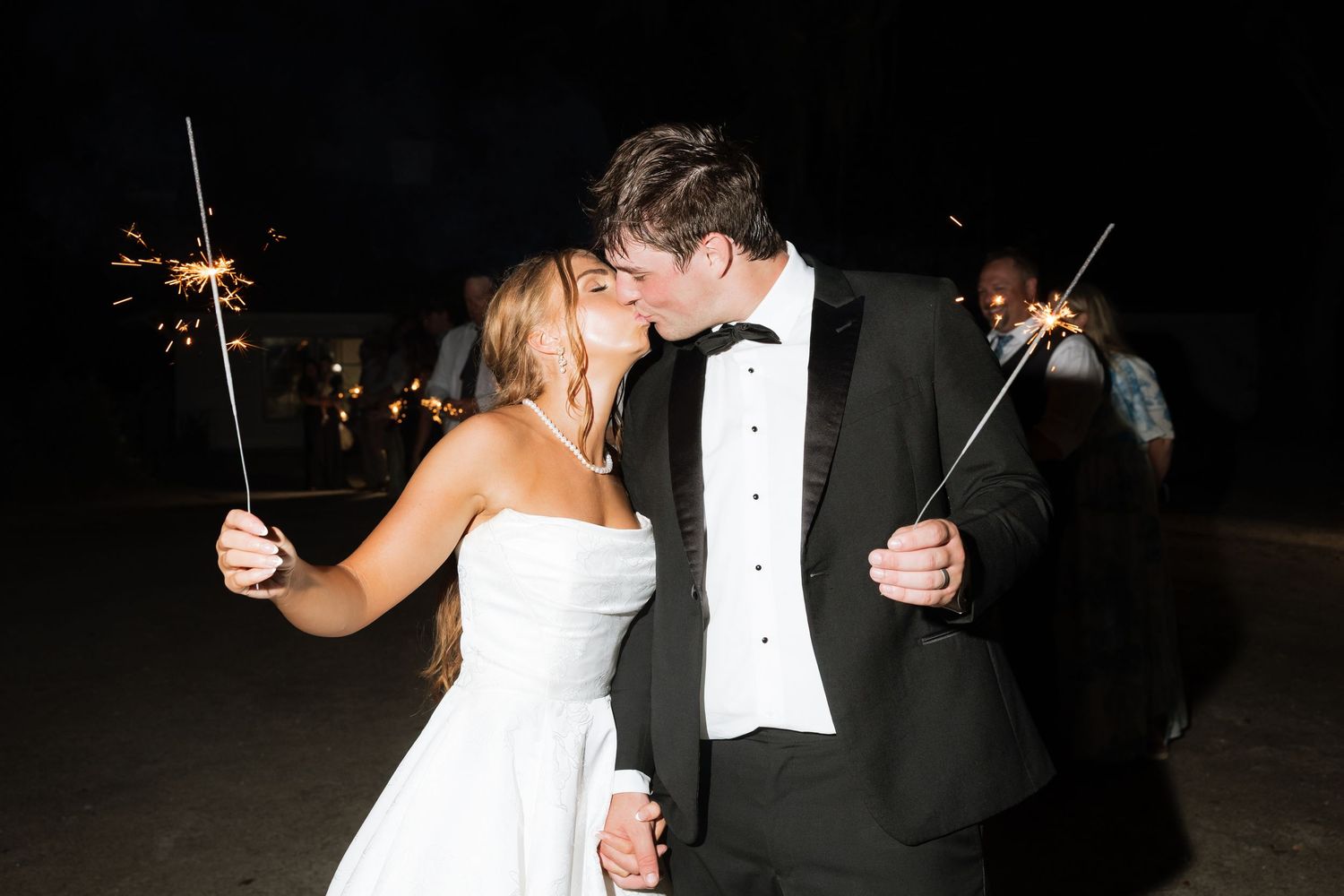A newlywed couple shares a romantic kiss while holding sparklers during their outdoor wedding reception.