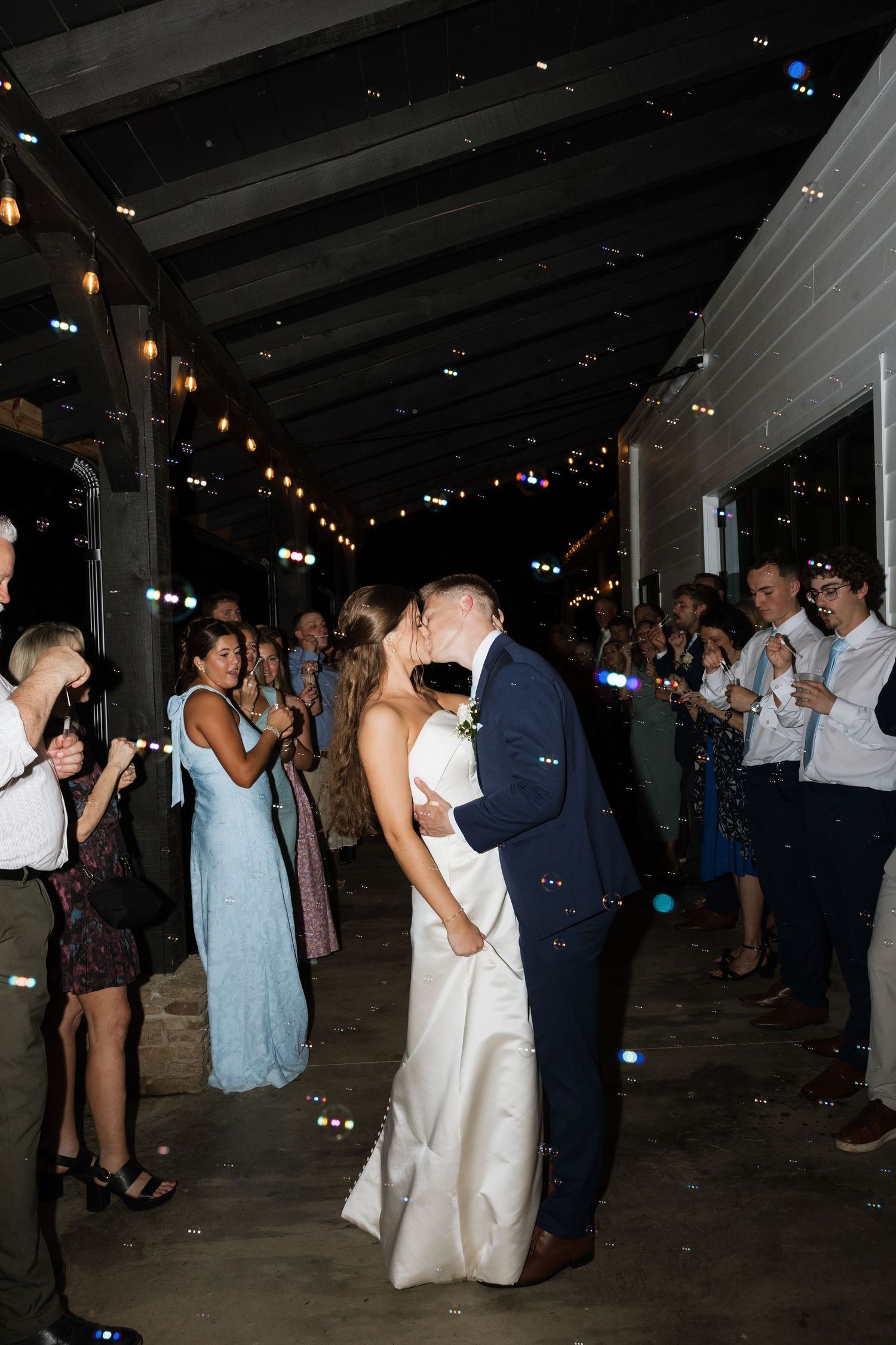 Bride and groom share a kiss on the dance floor surrounded by cheering guests during wedding reception at night.