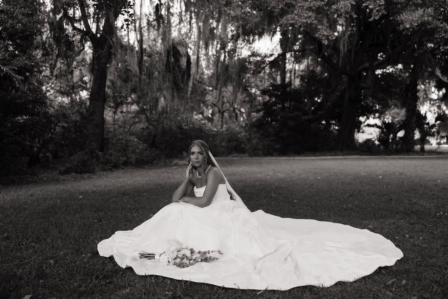 Black and white photo of a bride in a flowing gown seated on grass surrounded by moss-draped trees.